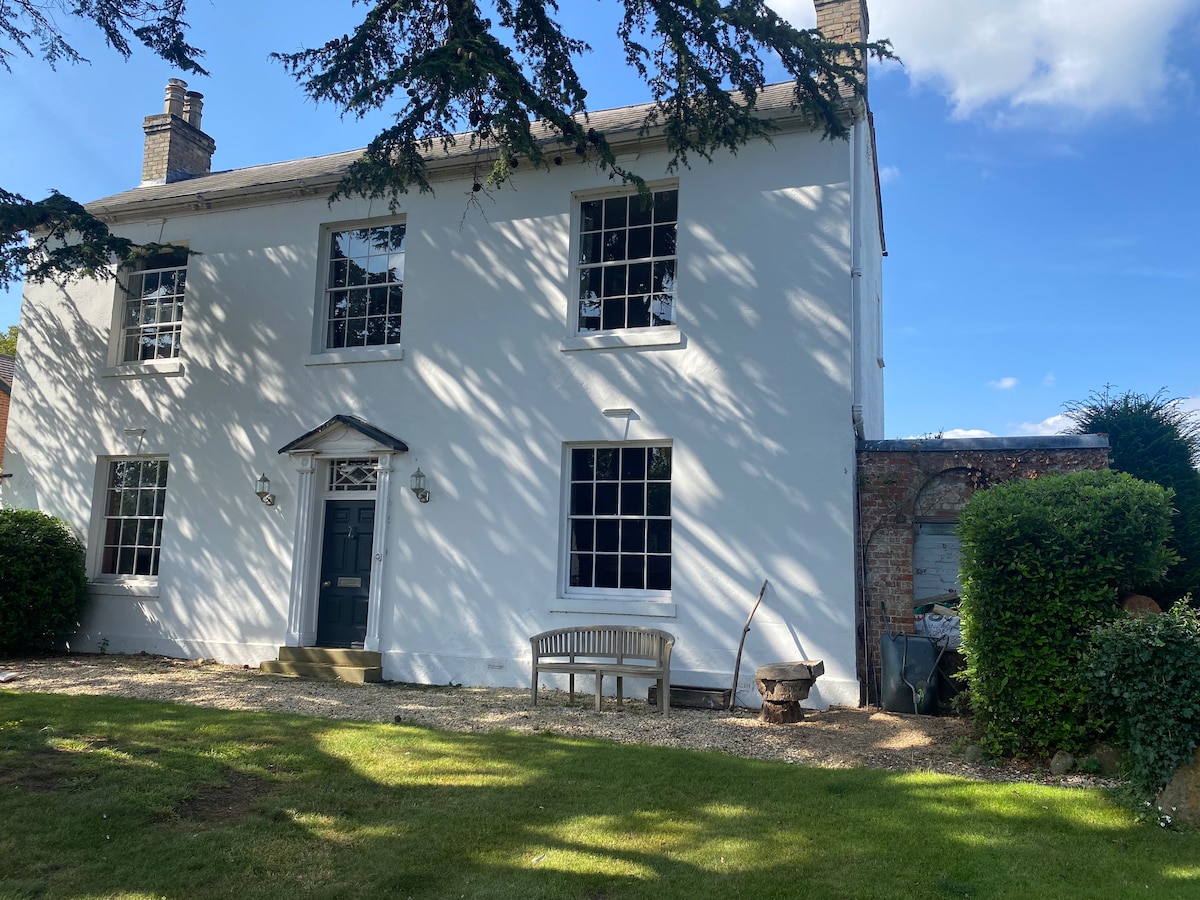 A classic two-story country home is presented, with a white exterior featuring large windows that allow natural light to fill the space. A wooden bench sits invitingly on a gravel pathway in front of the house, surrounded by manicured grass and greenery.