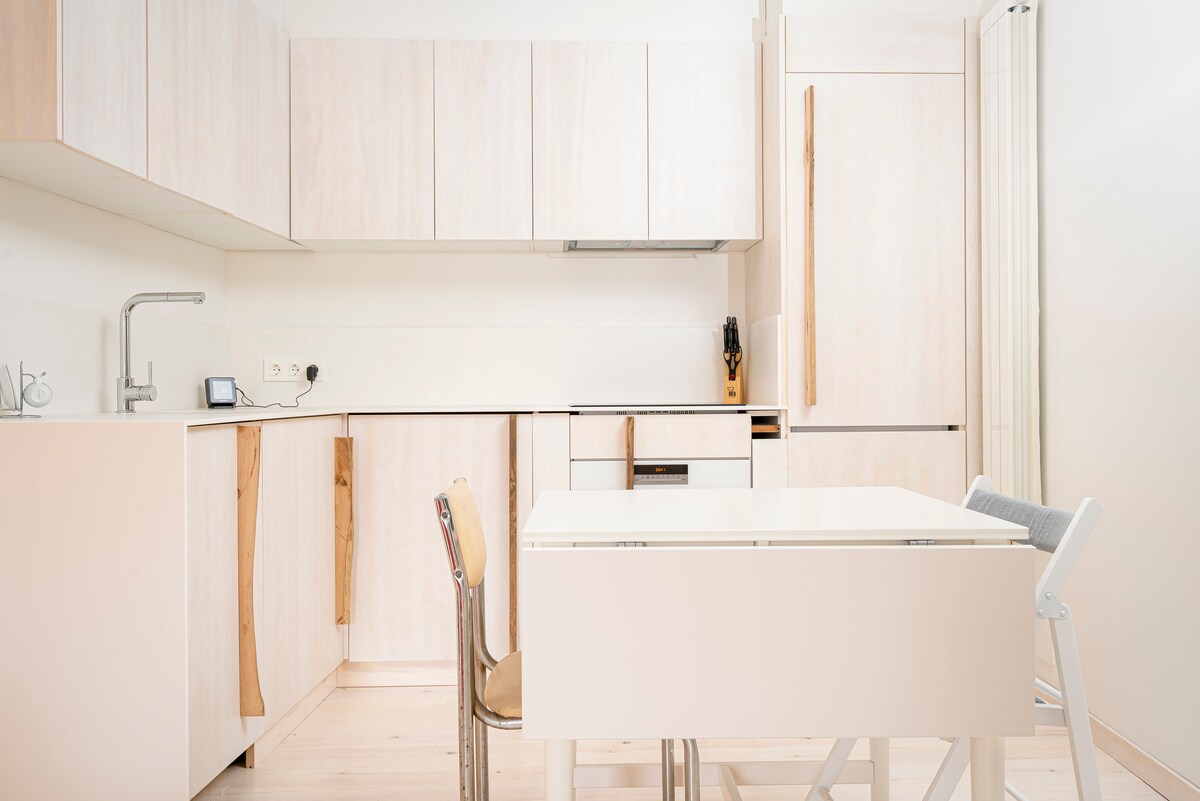 A modern kitchen space features light-colored cabinetry with wooden accents. A compact dining table is positioned in the center, accompanied by a mix of chairs. Countertops are clear, while appliances are integrated into the cabinetry, providing a streamlined appearance.