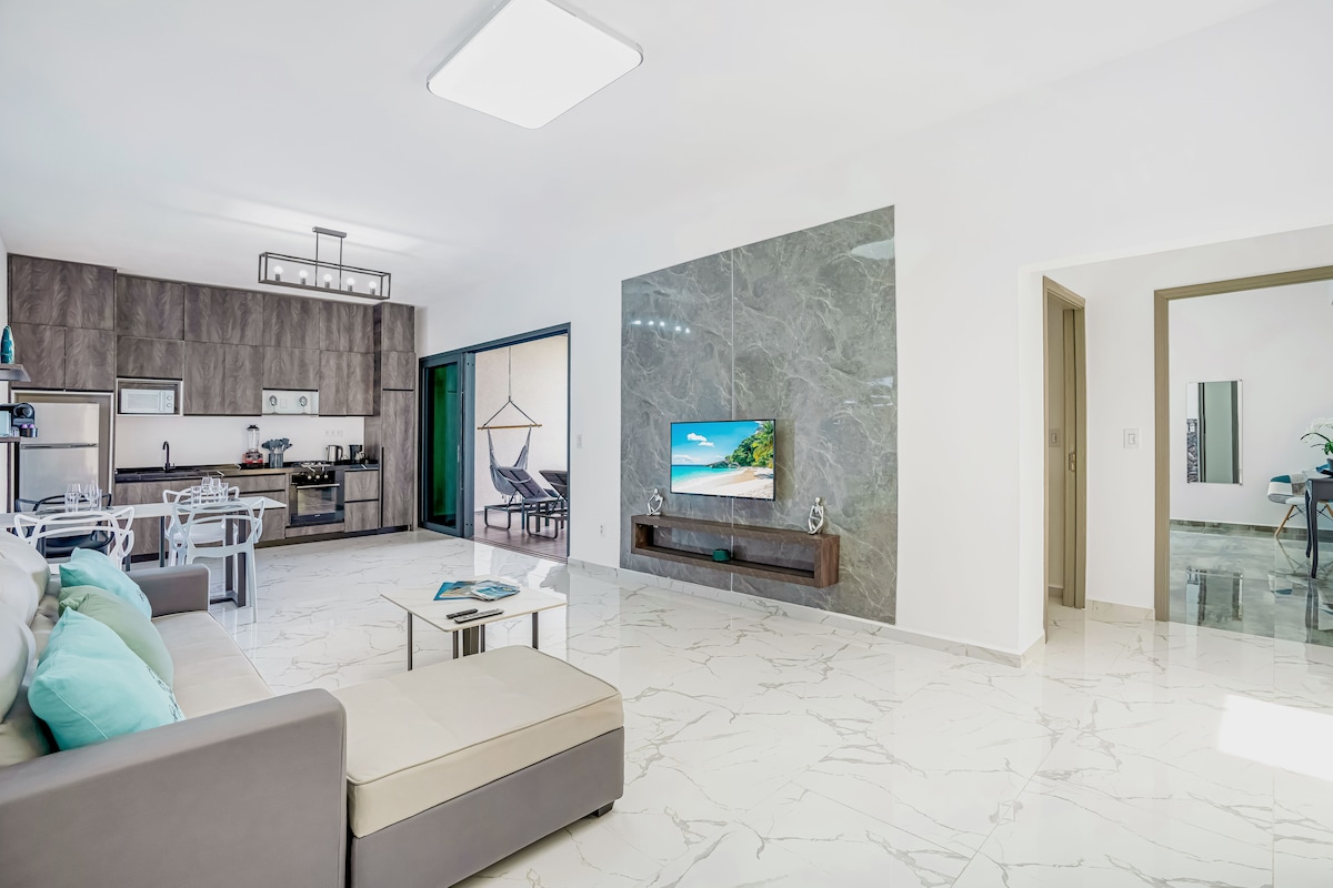 An open living area is presented, featuring a modern sofa with cushions and a coffee table. A wall-mounted TV is visible, displaying a tropical scene. The adjacent kitchen showcases sleek cabinetry, and sliding glass doors lead to a terrace with outdoor seating.