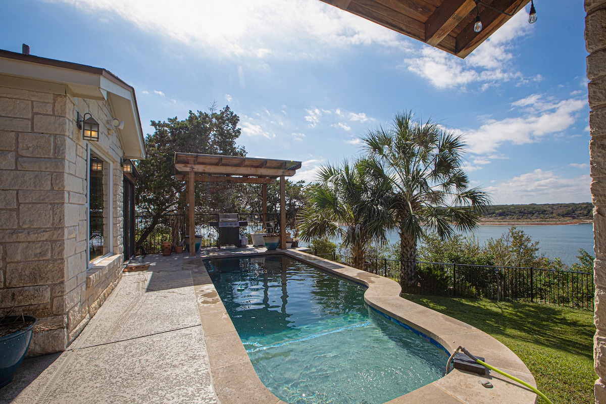 A serene outdoor area features a private pool surrounded by lush greenery. A pergola provides shade over a gas grill, with views overlooking the lake. Smooth stone and concrete surfaces offer easy access to the water and dining spaces.