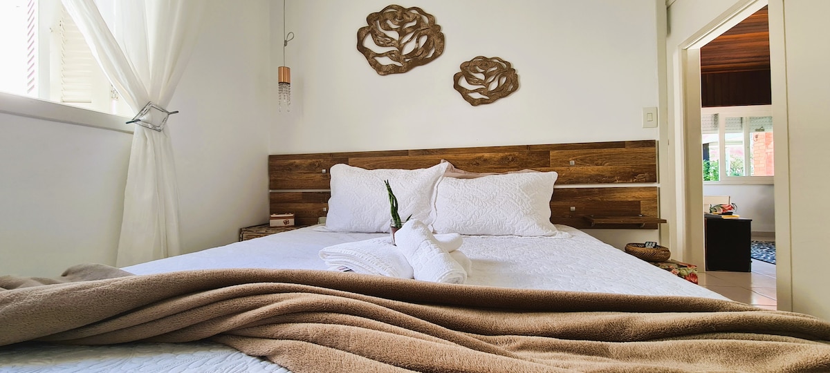 A spacious sleeping area features a wooden headboard with decorative wall art above the bed. Soft white linens are complemented by a neutral-toned blanket and rolled towels. Natural light filters through the curtains, illuminating the room's calming ambiance.