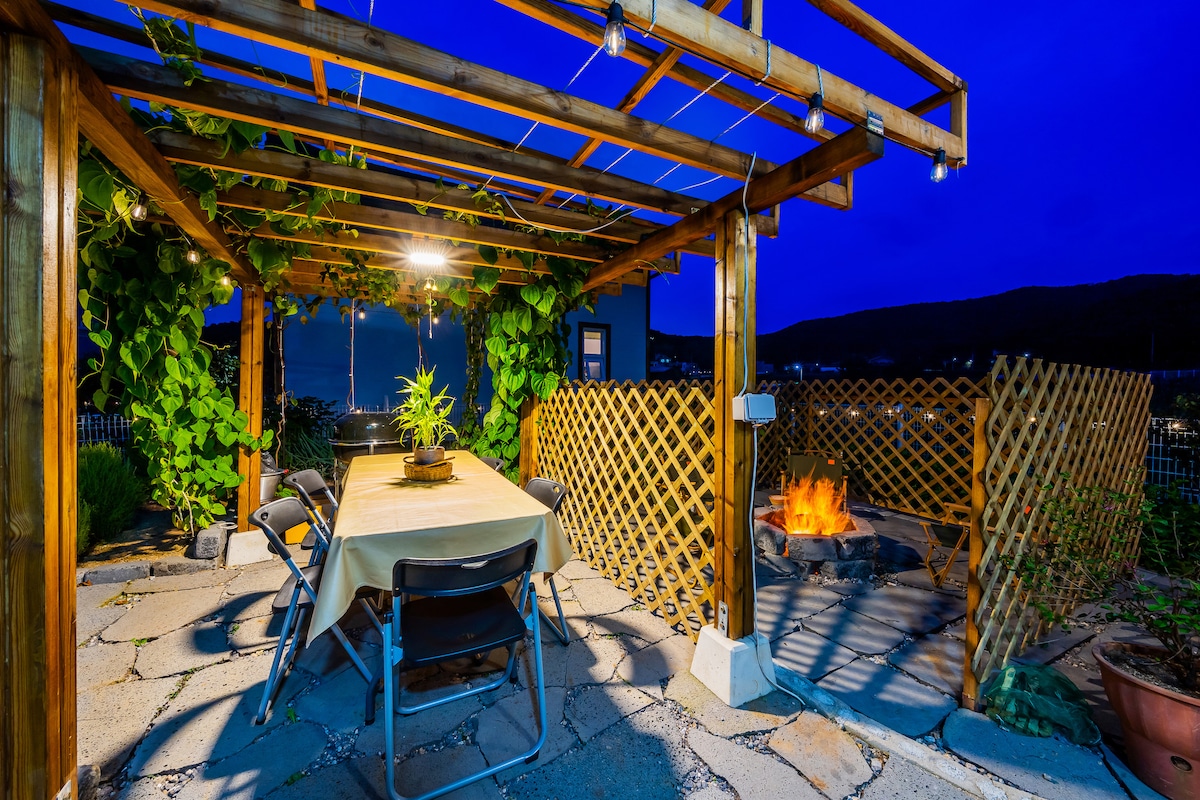 An outdoor seating area is featured under a wooden pergola adorned with climbing vines. A rectangular table is surrounded by chairs, and a fire pit is visible nearby, providing a cozy atmosphere in the evening light. Decorative string lights enhance the setting.