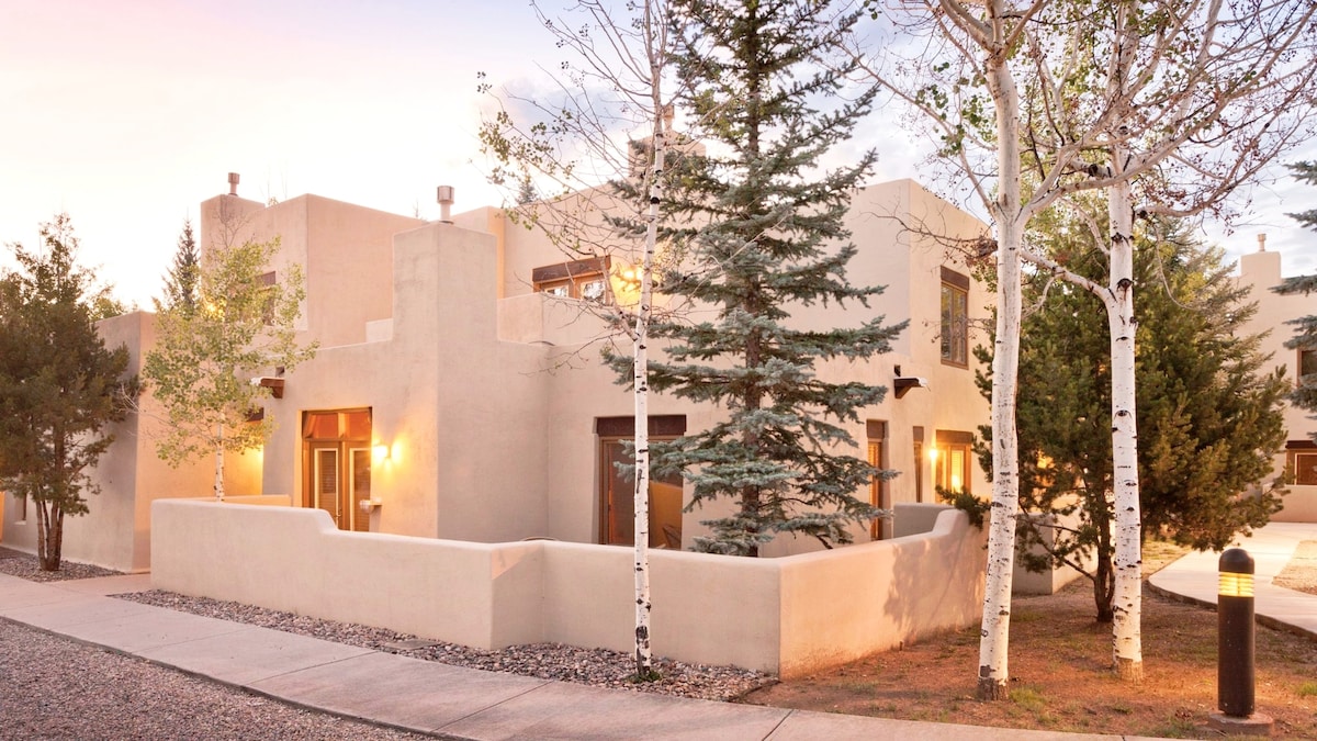 A modern adobe-style building is surrounded by trees and shrubs, showcasing a blend of natural and architectural elements. Soft lighting emanates from the windows, enhancing the warm tones of the stucco exterior during twilight.