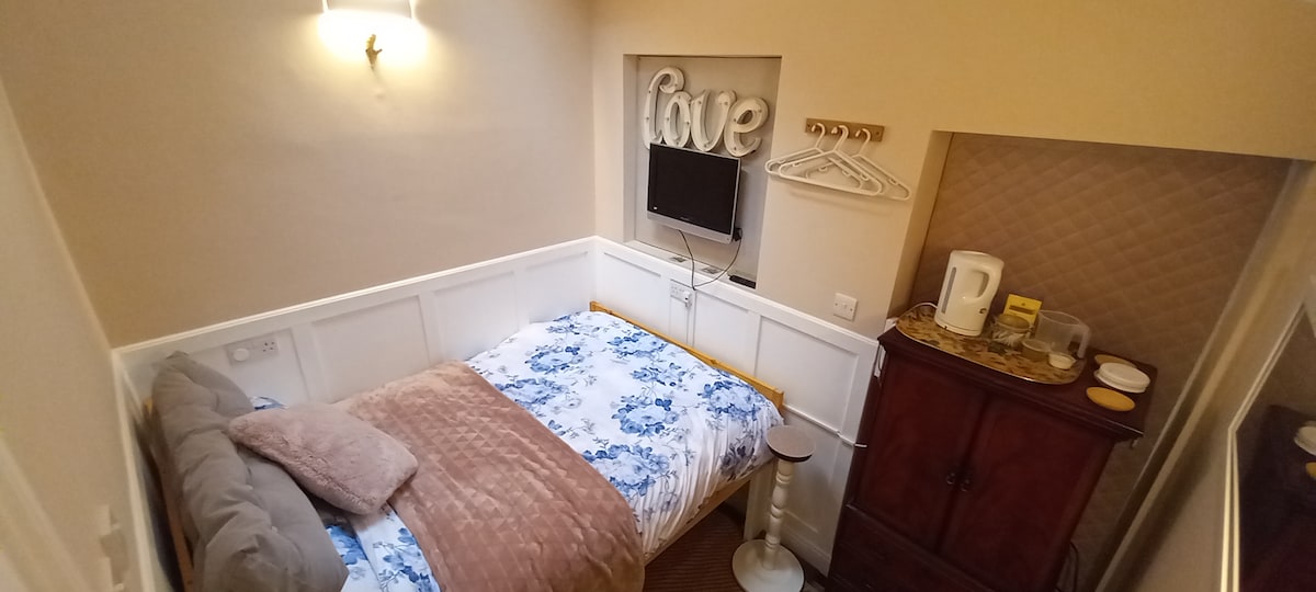 A cozy sleeping area featuring a queen medium firm mattress with a floral duvet. A slim television is mounted on the wall above a small cabinet, which holds tea-making facilities. A window adorned with decorative letters offers light to the room.