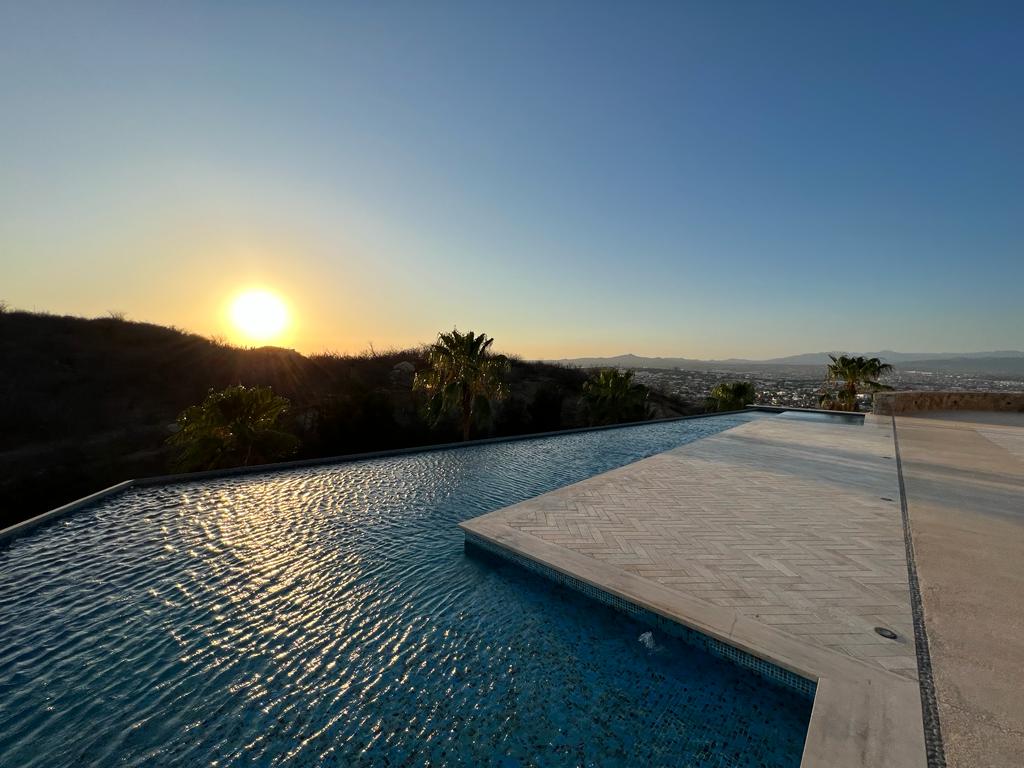 The infinity edge pool reflects the warm colors of the setting sun, creating a serene atmosphere. Lush palm trees frame the pool area, while the distant city and mountains provide a picturesque backdrop against the clear evening sky.