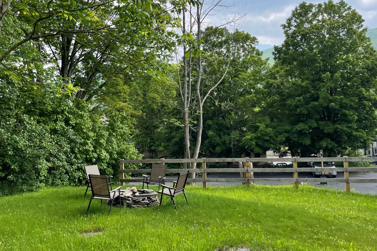 A shared yard features a fire pit surrounded by four chairs on a lush green lawn. Tall trees and shrubs provide natural privacy and shade. A wooden fence lines the area, enhancing the tranquil outdoor space.