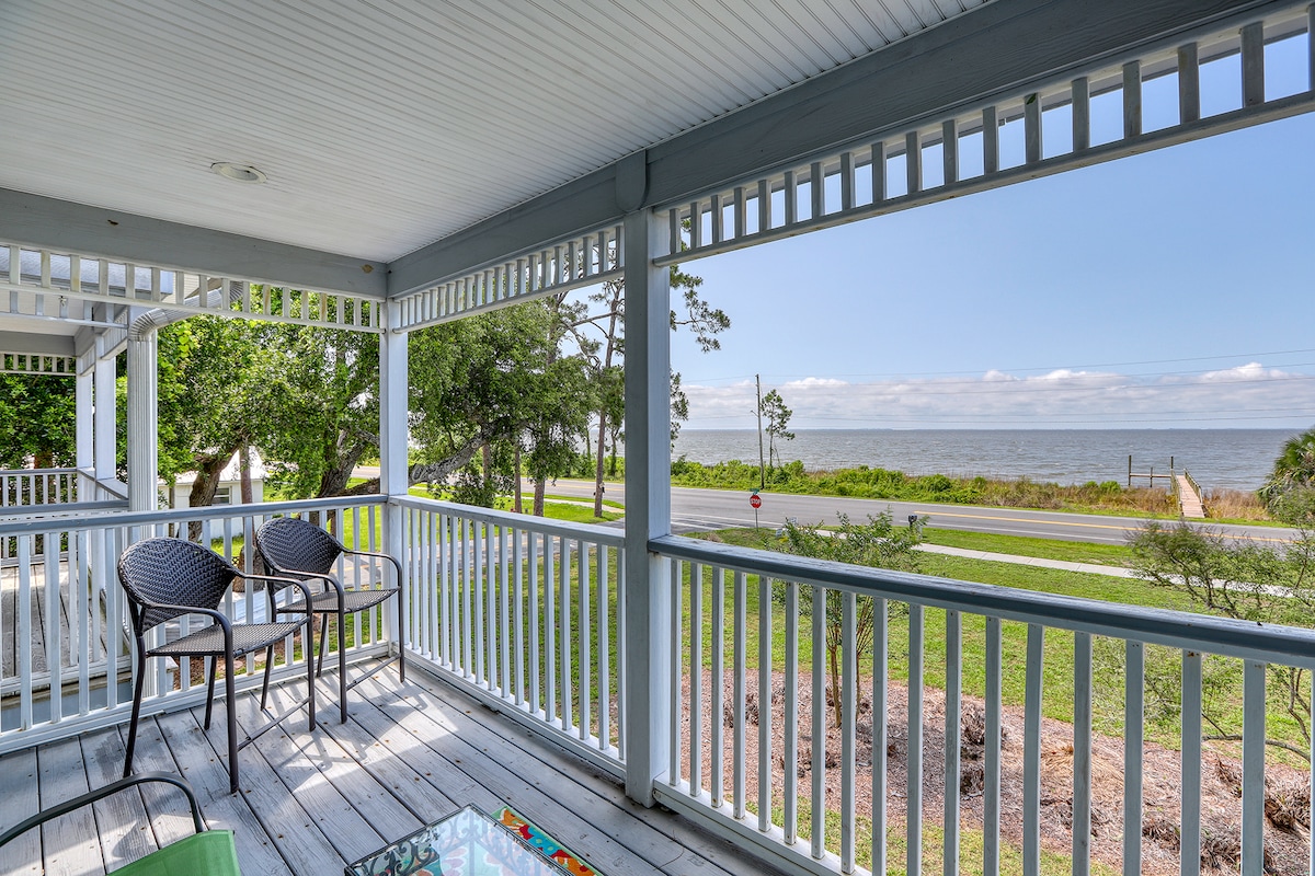 A covered porch is shown with a wooden deck and white railings. Two chairs are placed beside a glass table, offering a view of the bay in the distance. Lush greenery and a road are visible beyond the porch, enhancing the serene setting.