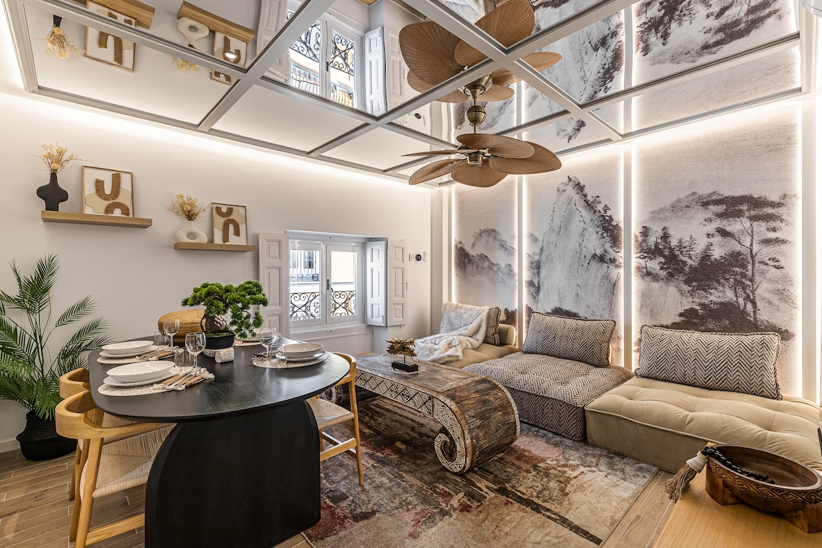 A spacious living area is adorned with stylized wall art featuring natural landscapes. A circular dining table set for four is positioned beside a comfortable seating area, which includes a sectional sofa and a decorative coffee table. Soft lighting creates a cozy ambiance.