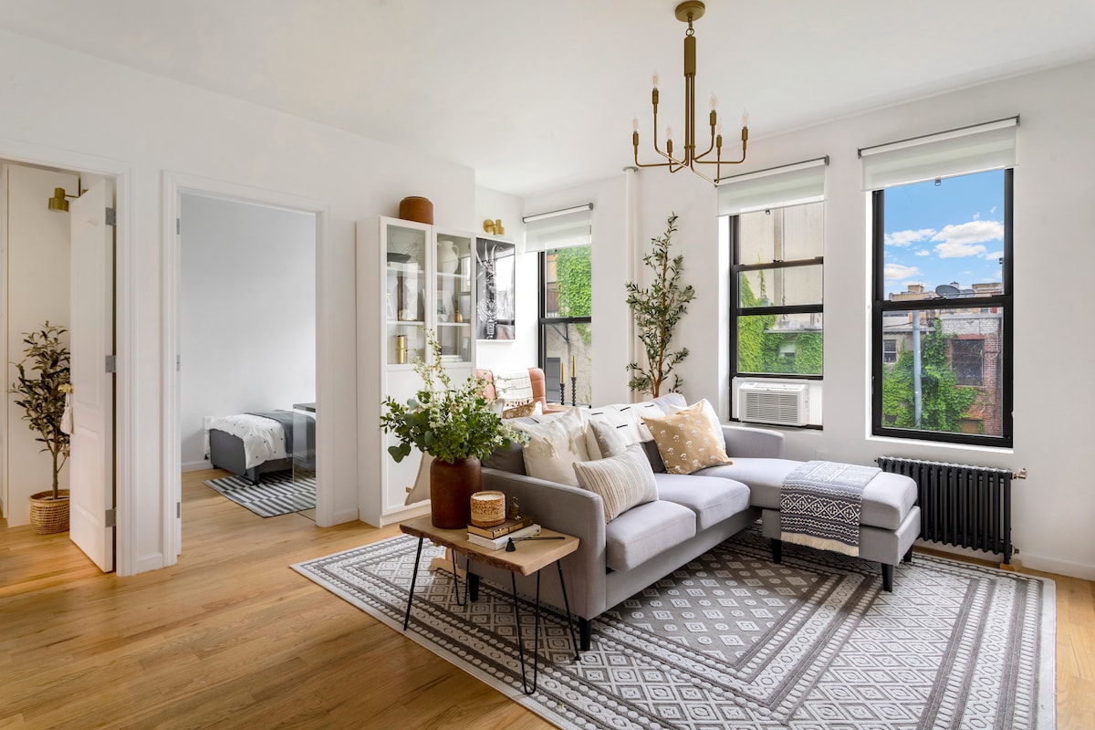 An inviting living area features a spacious gray sectional sofa adorned with decorative throw pillows. Natural light streams through large windows, illuminating a stylish area rug and a coffee table with floral arrangements. A glimpse of a cozy bedroom is visible through an open doorway.