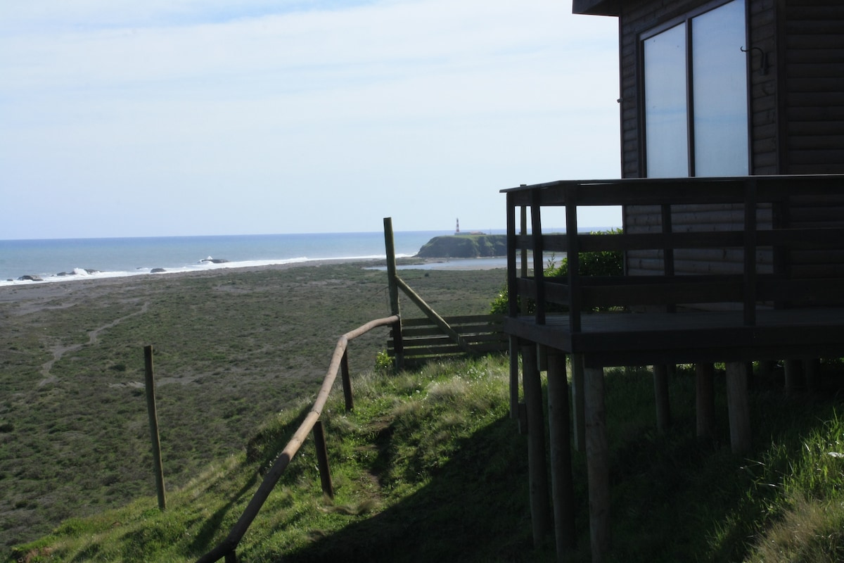 The image captures a wooden structure overlooking a lush green landscape leading to the beach. A wooden deck extends from the house, providing views of the ocean and a distant lighthouse. Gentle waves can be seen along the coastline under a clear blue sky.