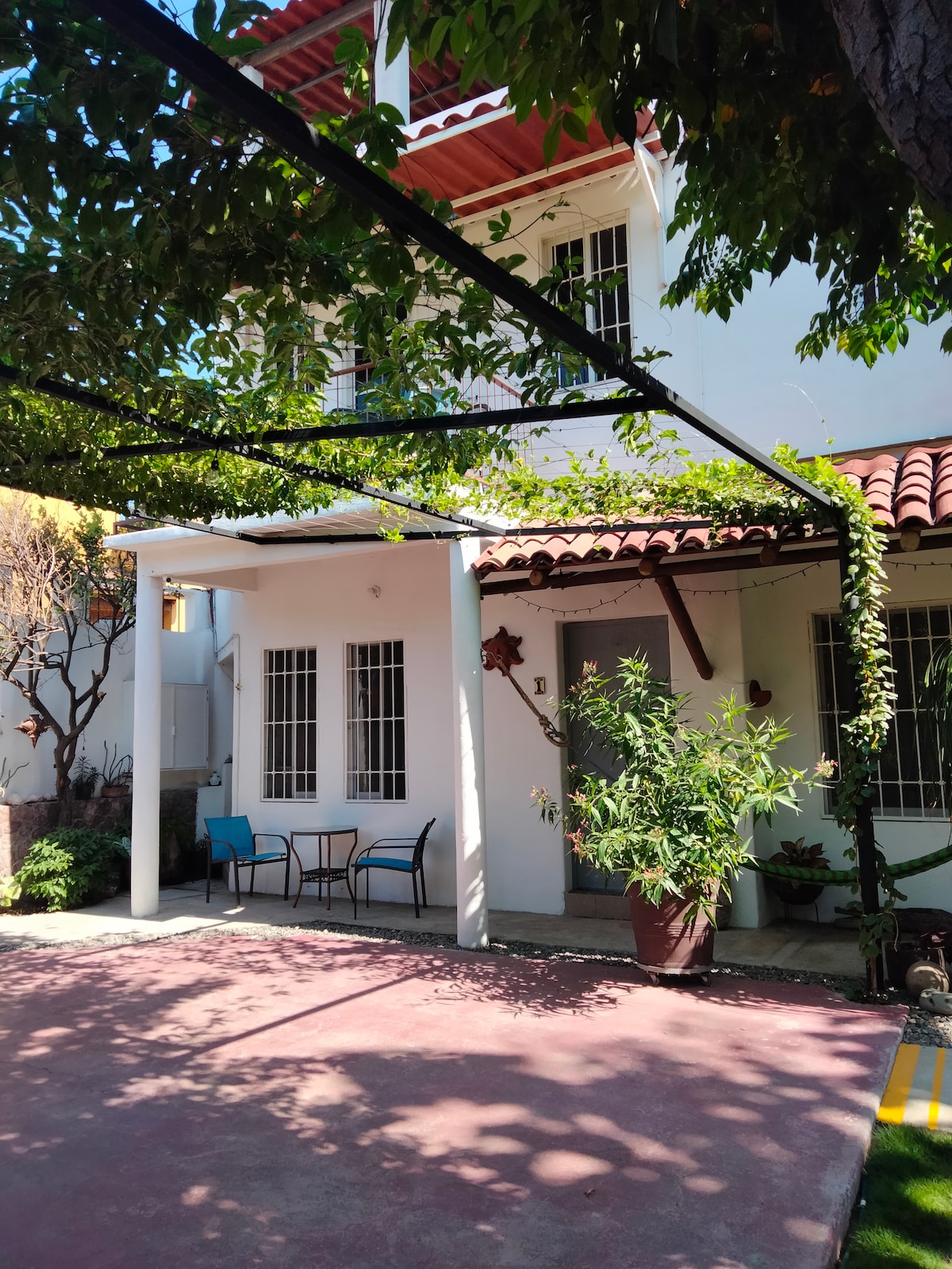 A shaded outdoor patio area is visible, featuring two chairs and a small table positioned under a trellis covered with greenery. The walls of the bungalow are adorned with white paint, and a colorful potted plant adds a touch of nature to the setting.