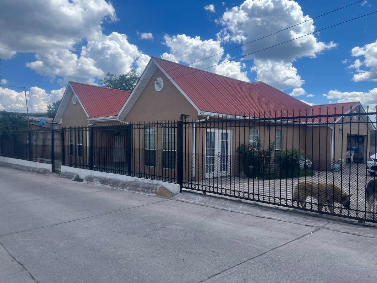 A single-story building is set behind a secure metal fence, featuring a red metal roof. Two prominent gable ends are visible, and large windows allow natural light to fill the interior. A gravel driveway is seen in front of the house, surrounded by greenery.
