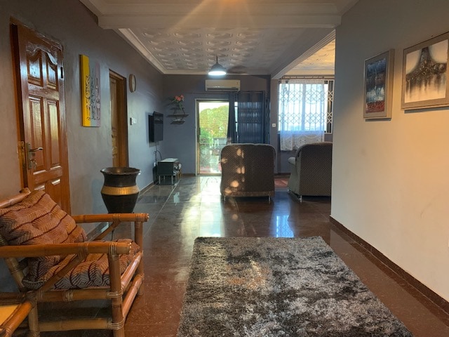 A spacious living area features a bamboo chair next to a decorative table. A large doorway leads to a balcony, while light filters through mesh curtains. Two cushioned chairs are arranged on a plush carpet, complemented by wall art and a television mounted on the wall.