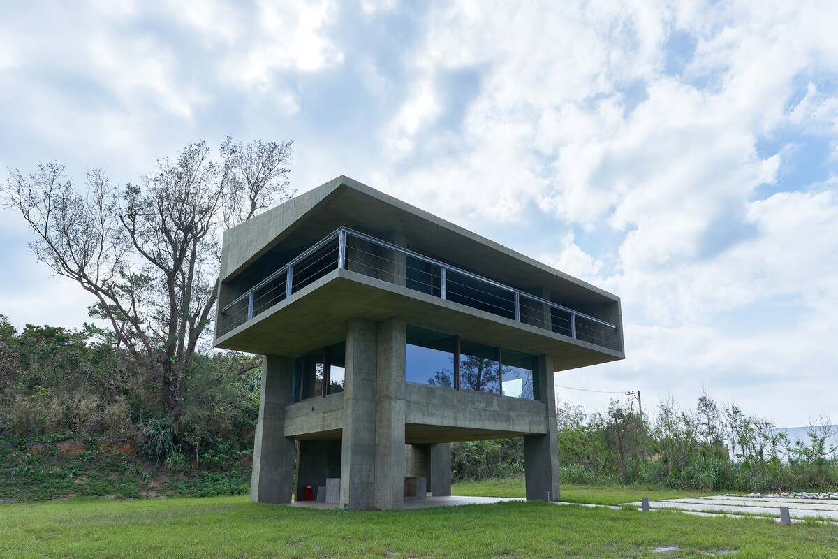 A modern two-story home features a concrete exterior with large glass windows. The structure stands elevated on sturdy columns, offering a sense of openness. The surrounding landscape includes greenery and a gradual slope, providing a serene backdrop under a partially cloudy sky.