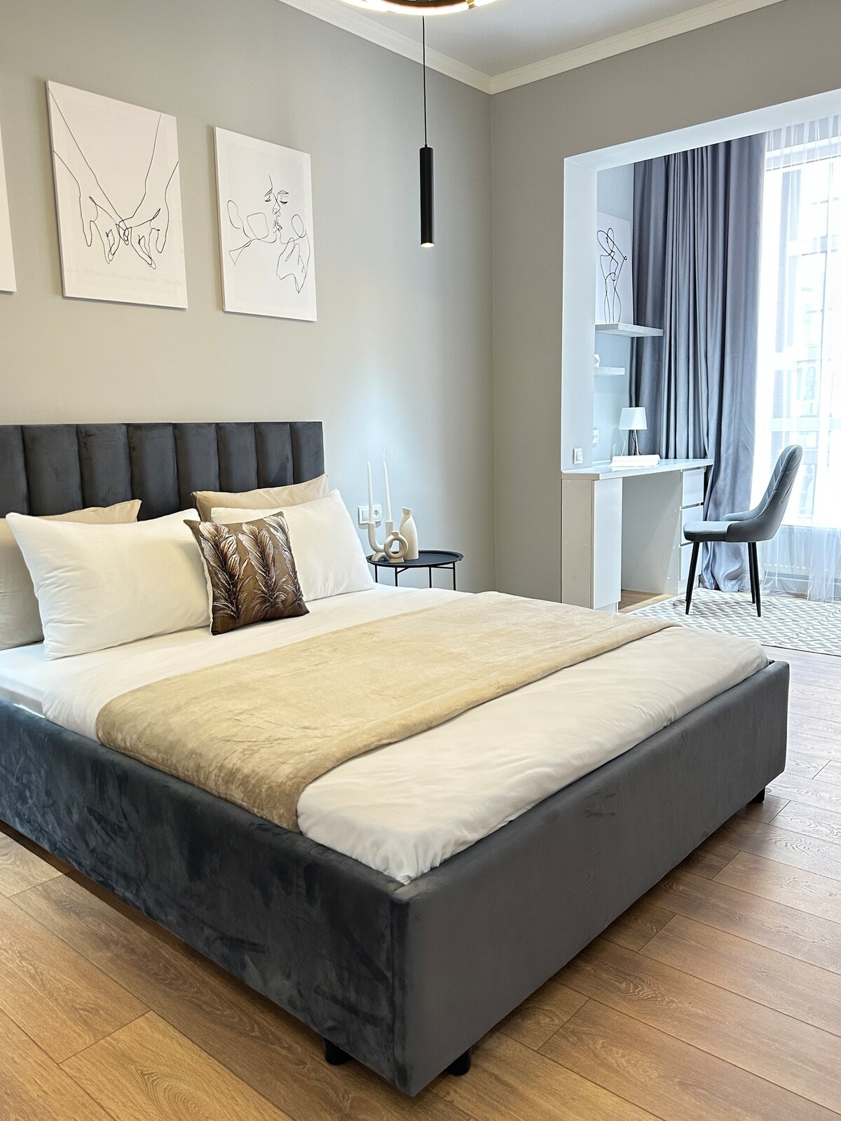 The bedroom features a spacious bed with a soft gray velvet frame, complemented by decorative pillows. Light pours in through a large window, while minimalistic artwork adorns the walls. A small workspace with a chair is situated next to the window, providing a cozy, functional area.