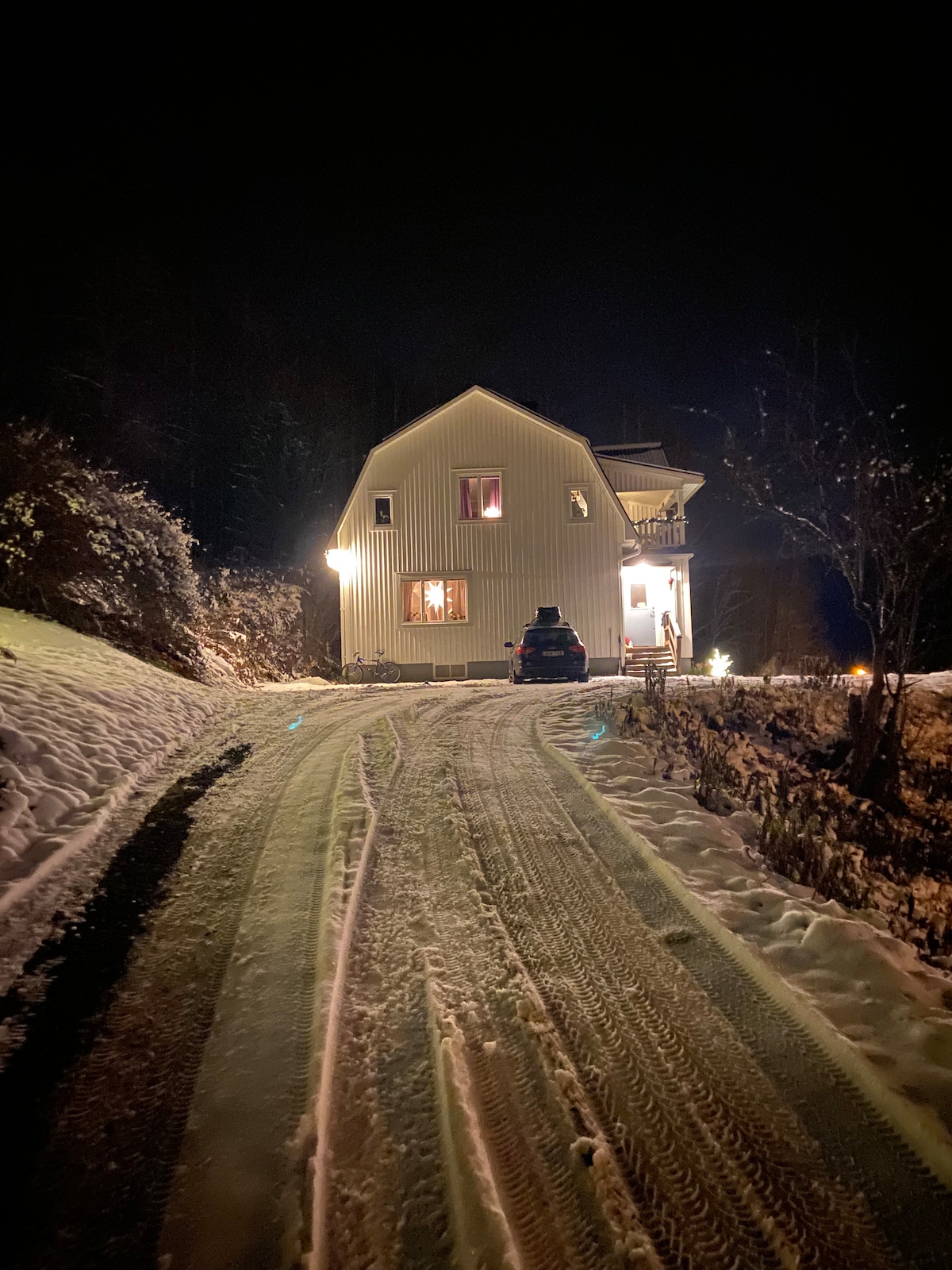 The house is viewed at night from a snowy driveway, with warm lights illuminating its exterior. A vehicle is parked in front, and the surrounding area features snow-covered ground and trees, creating a serene winter scene.