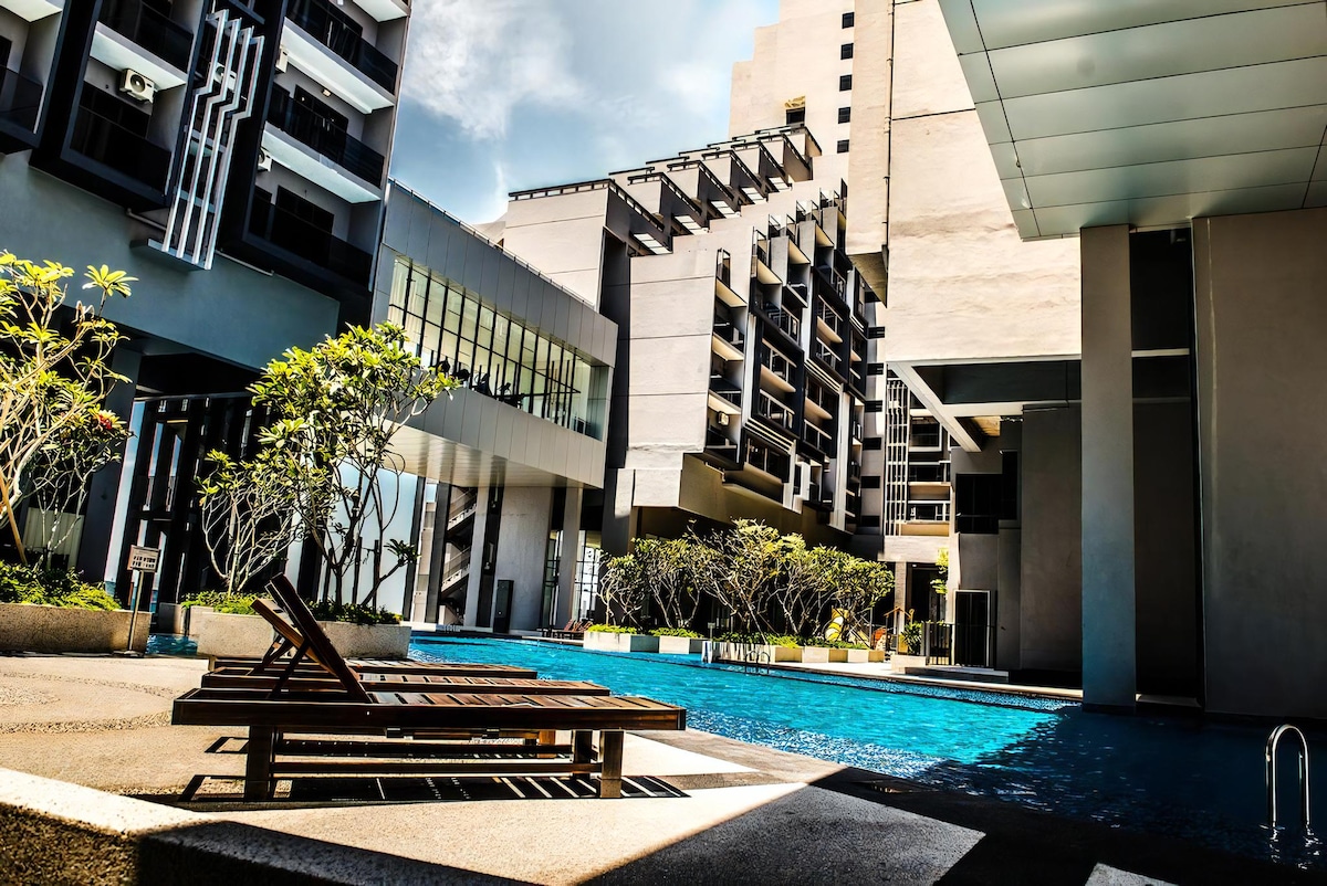 A tranquil outdoor area features a swimming pool surrounded by modern buildings. Wooden lounge chairs are positioned beside the pool, while lush greenery is present in landscaped planters. The scene is complemented by clear skies, enhancing the inviting atmosphere of the space.