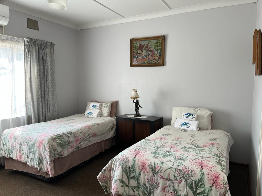 A room features two twin beds dressed in floral bedding, positioned side by side. A bedside table with a lamp stands between them. Light enters through a large window, adorned with sheer curtains, illuminating the artwork hanging on the wall above.