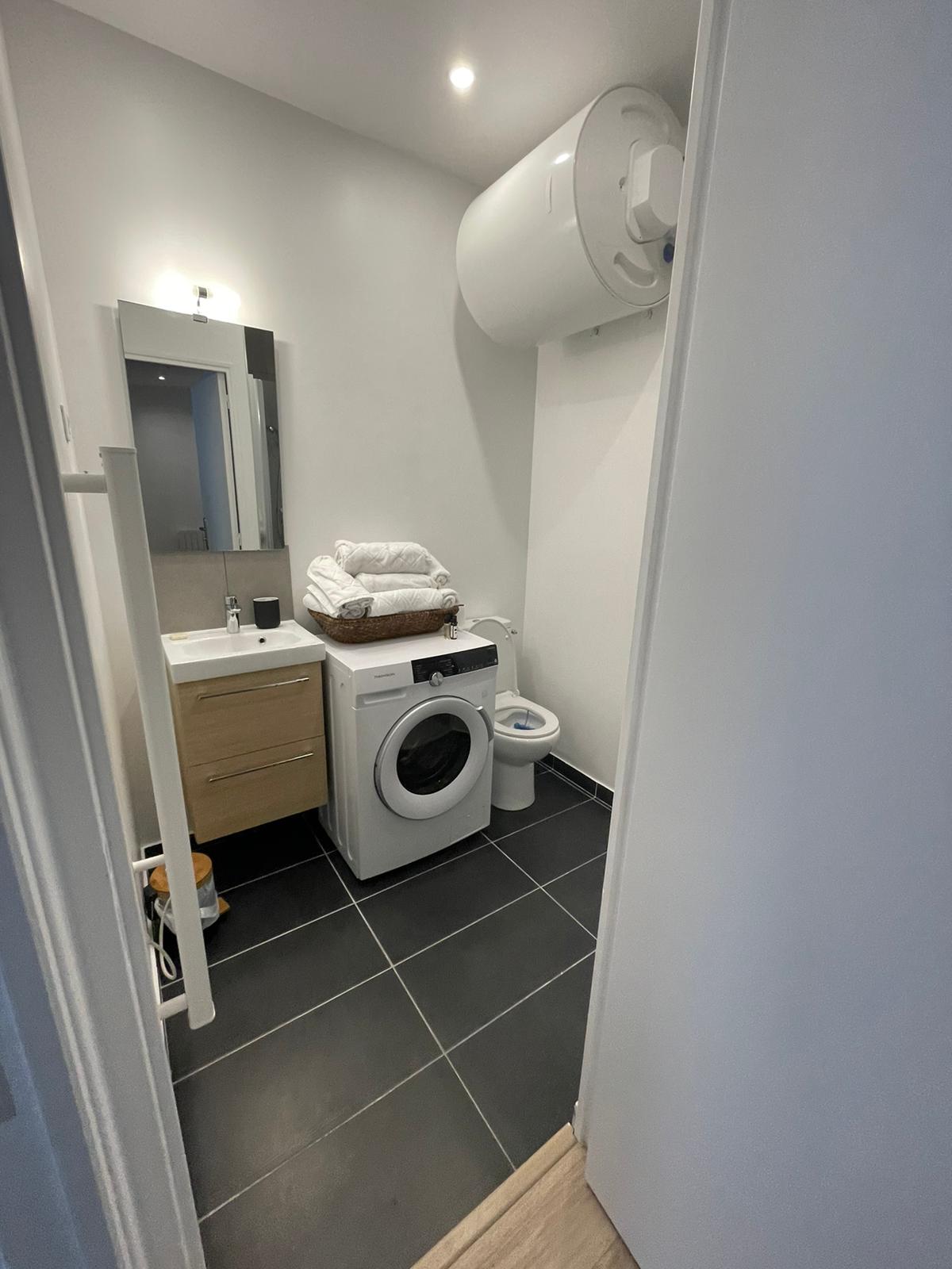 The bathroom features a modern design with dark tile flooring. A washing machine is placed next to a compact vanity unit with a mirror. Fresh towels are neatly arranged on top of the machine. A toilet is also present, with ample light provided by ceiling fixtures.