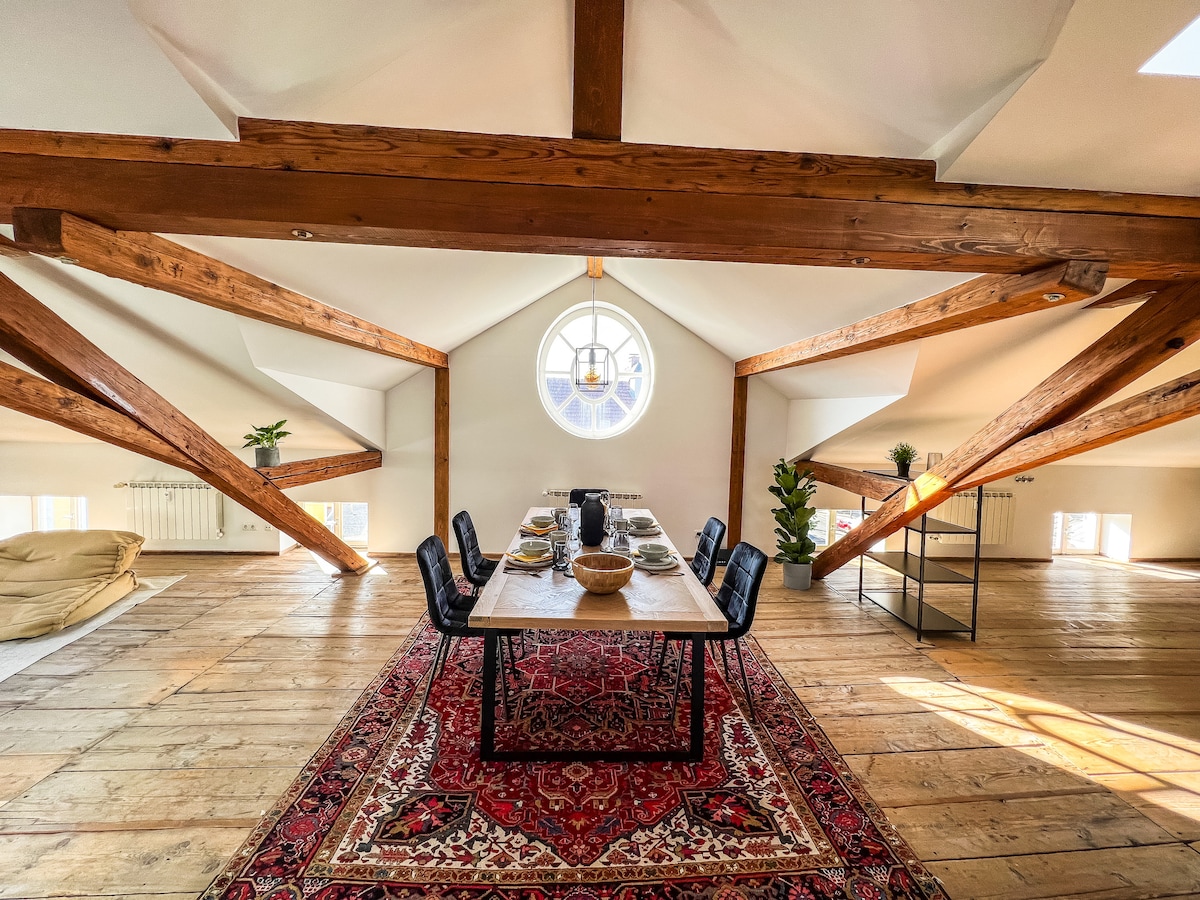 An airy dining area features a long table set for a meal, surrounded by black dining chairs. Large circular windows allow natural light to fill the space, illuminating the wooden beams overhead and the patterned rug beneath. A cozy seating area is visible in the background.