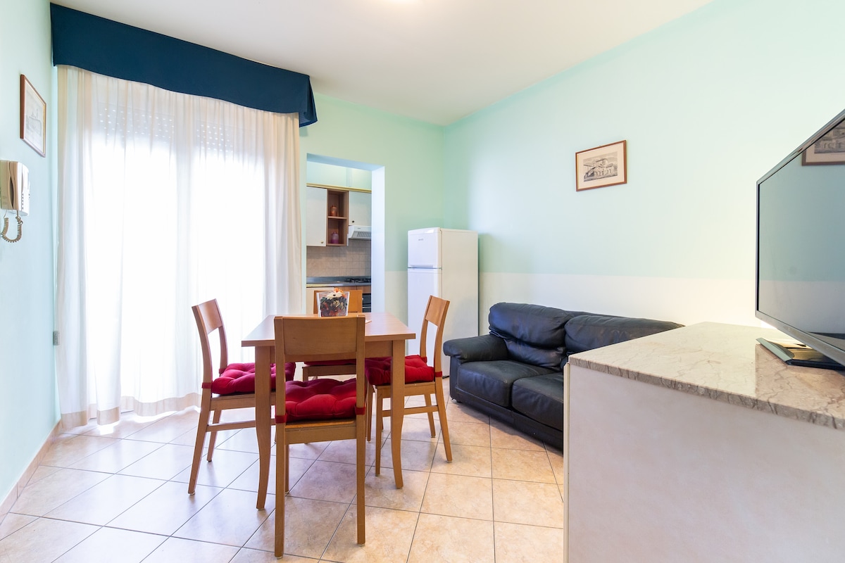 A dining area features a wooden table surrounded by four chairs with red cushions. A dark sofa is positioned nearby, and a bright window allows natural light to fill the space. The kitchenette is partially visible in the background, along with a white refrigerator.