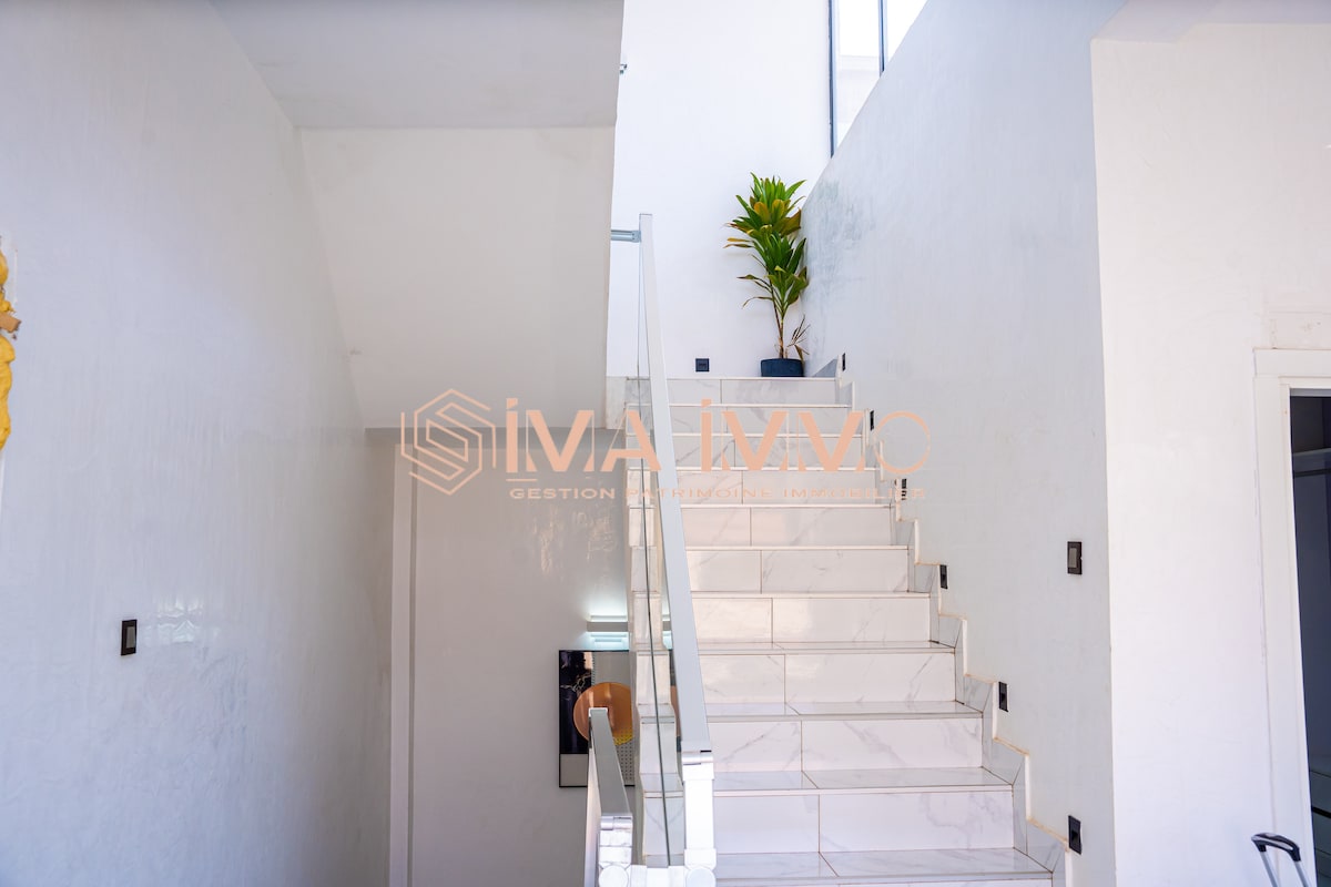 A well-lit staircase ascends with white marble tiles, featuring a sleek glass railing. A small potted plant in a corner adds a touch of greenery against the bright walls.