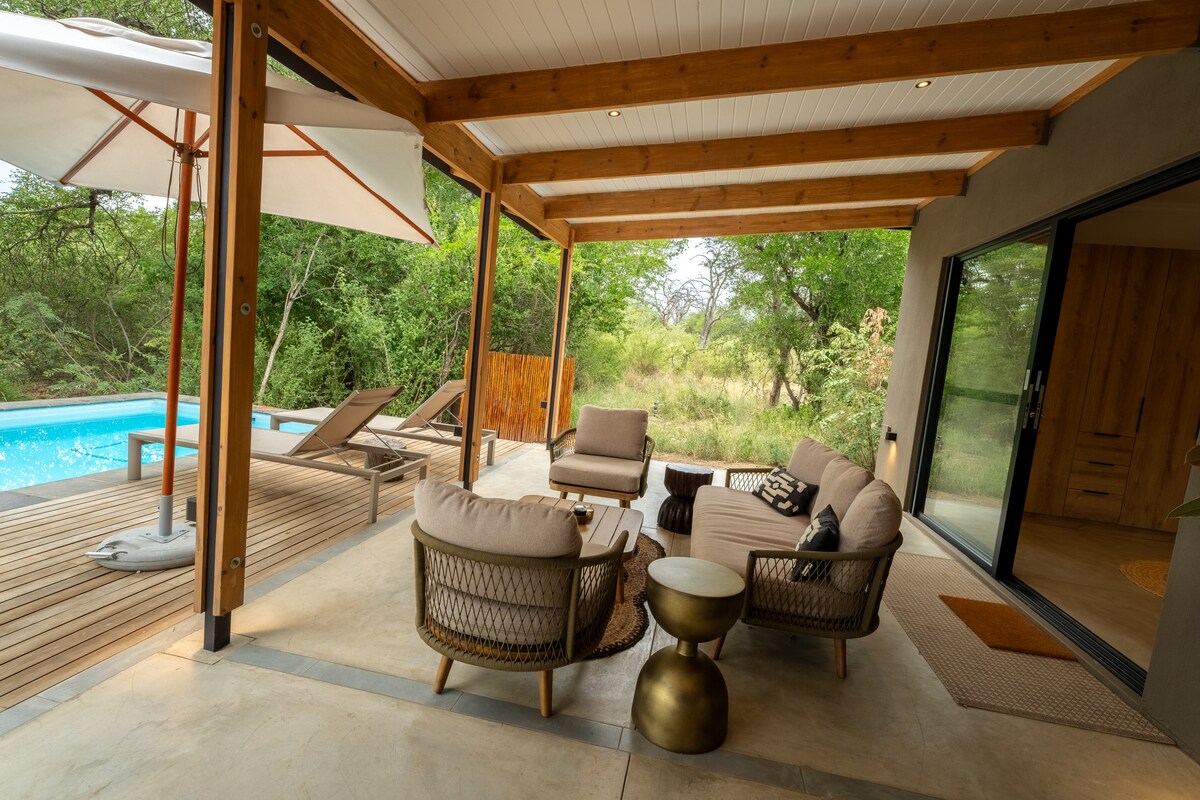 A covered patio is furnished with comfortable seating, featuring soft cushions and accent tables. A heated pool is seen nearby, surrounded by lounge chairs. Lush greenery is visible in the background, enhancing the natural setting.