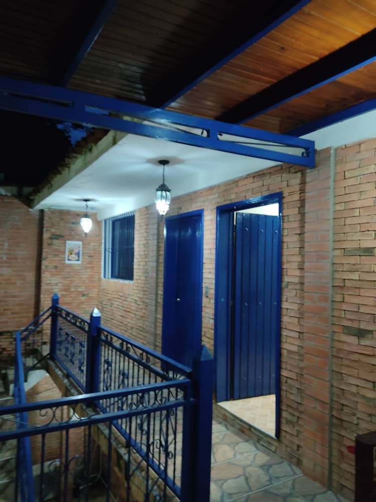 The entrance area is defined by brick walls and a wooden ceiling, complemented by lantern-style lighting. A blue railing frames the walkway, leading to a solid blue door. The two windows are set with protective grills, enhancing both security and visibility.