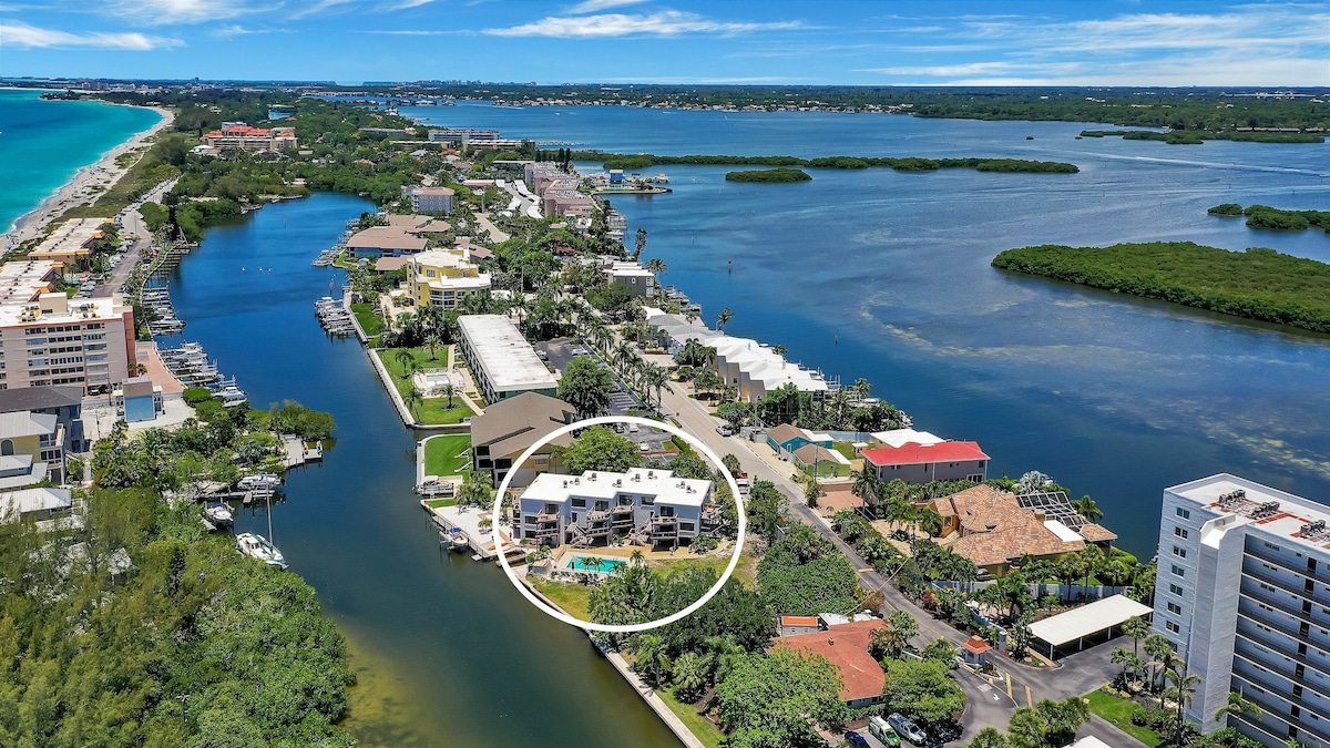 An aerial view showcases the waterfront property, highlighting the building's proximity to the canal. Nearby greenery and water feature prominently, while the surrounding area includes various residential and commercial structures, indicating a vibrant community. The pool is visible, situated on the property.