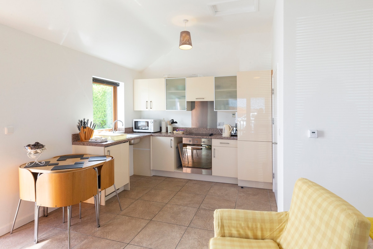 A modern kitchen features sleek cabinetry and a light granite countertop. Essential appliances such as a microwave and electric oven are visible. A round dining table with a patterned top complements a yellow accent chair positioned nearby. Natural light streams in through a window.