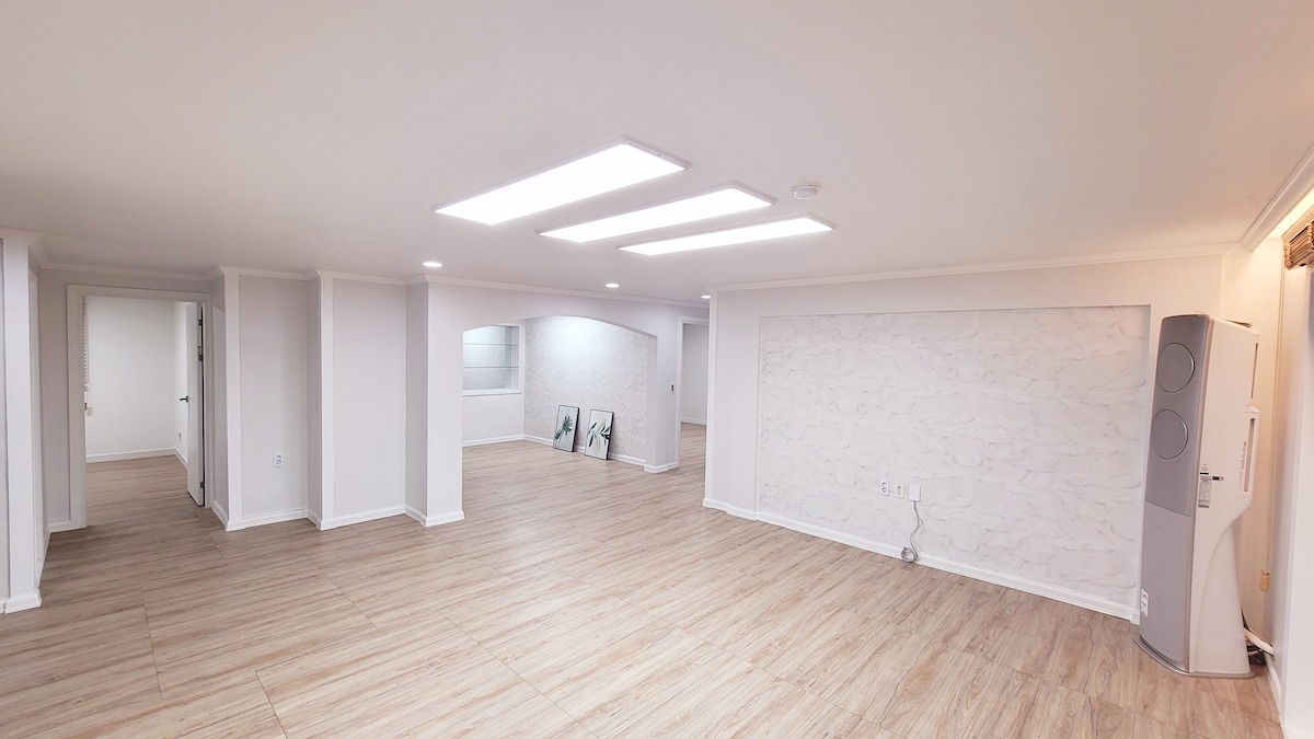 A spacious living area features a light-colored wooden floor and is illuminated by overhead lights. The walls are painted in neutral tones with a textured feature wall. Open spaces create a sense of flow, with distinct sections visible that can accommodate various furniture arrangements.