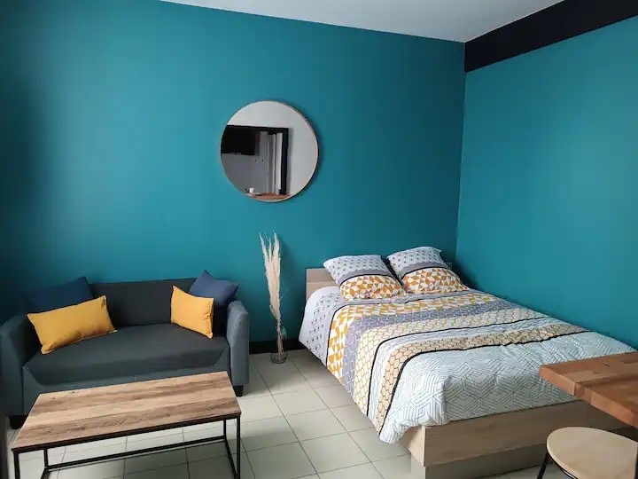 A studio space features a double bed with a patterned comforter and decorative pillows. A gray sofa with yellow accents complements a wooden coffee table. A round mirror hangs on the vibrant teal wall, enhancing the room's lighting. The flooring is tiled, contributing to the overall decor.