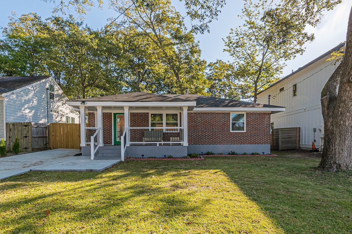 A renovated brick home is presented with a welcoming front porch and two chairs positioned for relaxation. The well-maintained lawn is bordered by diverse trees and shrubs. Sunlight filters through the branches, creating a pleasant outdoor space in the established neighborhood.