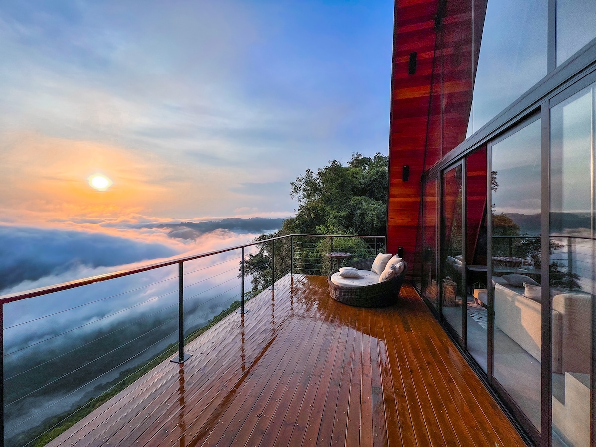 A spacious balcony features wooden flooring and modern seating, offering stunning views of the sunrise above a sea of clouds. Expansive glass walls enhance the connection with nature, allowing for unobstructed sightlines to the surrounding landscape.
