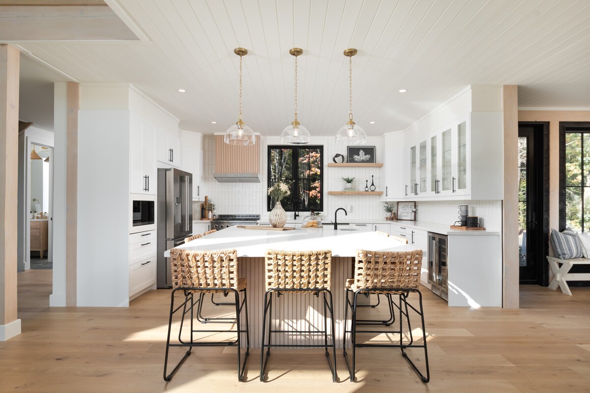 The modern kitchen features a large central island with four woven bar stools. Crisp white cabinetry is complemented by stainless steel appliances and light wood accents. Three pendant lights hang above the island, illuminating the space where natural light filters in through large windows.