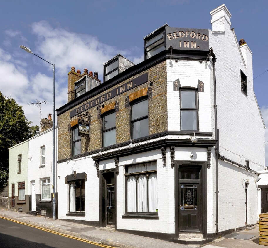 The exterior of the Bedford Inn is presented, showcasing a blend of brick and white façades. The establishment features large, framed windows and a prominent sign above the entrance. Surrounding greenery and a clear sky contribute to the inviting appearance of the pub.