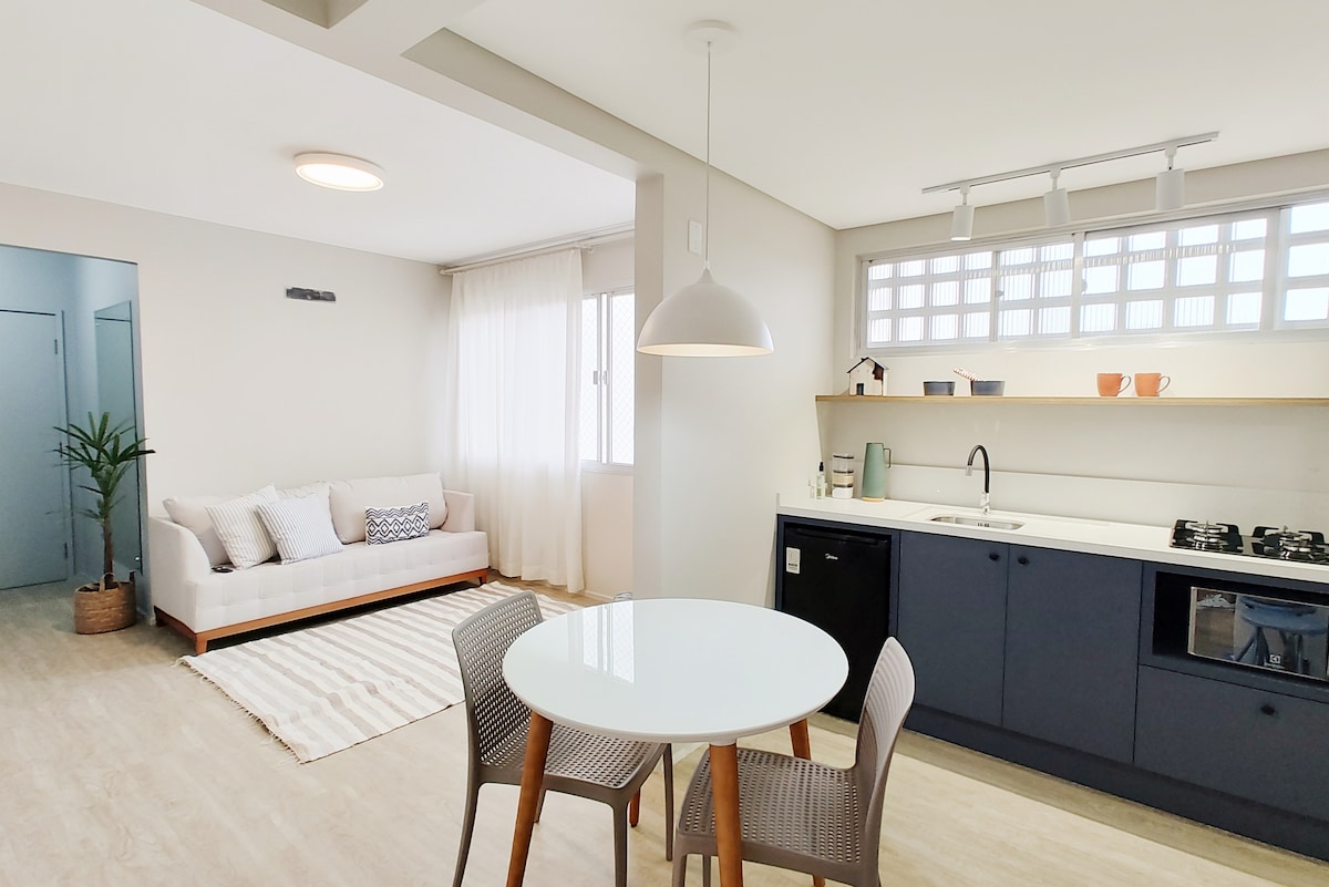 A bright and airy living space features a cozy seating area with a light-colored couch and a round dining table with four chairs. A compact kitchen is visible, equipped with modern appliances and a wooden shelf displaying decorative items.