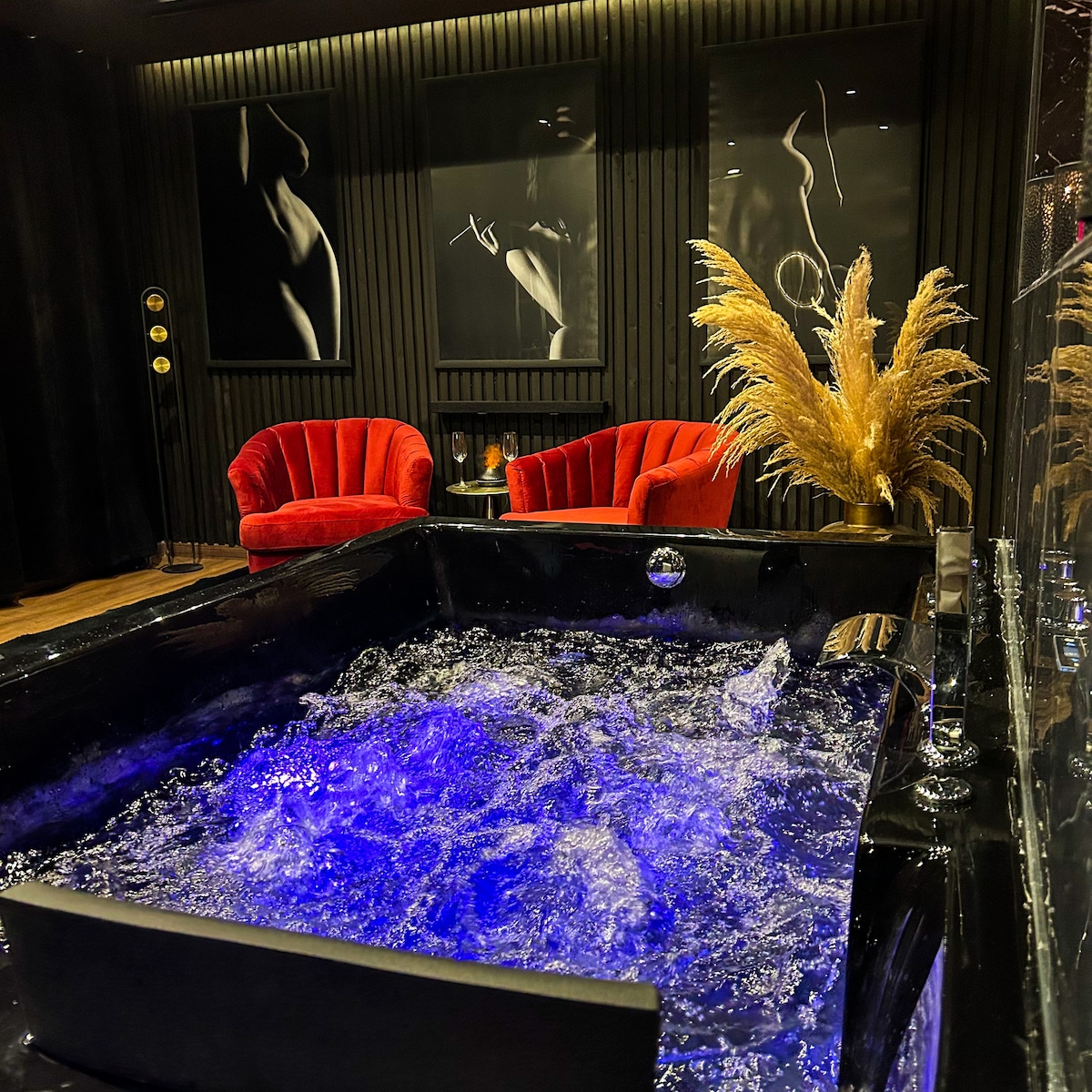 A modern jacuzzi is set against dark textured walls, with blue lighting illuminating the bubbling water. Two plush red chairs are positioned nearby, enhancing the intimate setting. Art pieces featuring abstract silhouettes are displayed on the adjacent wall.