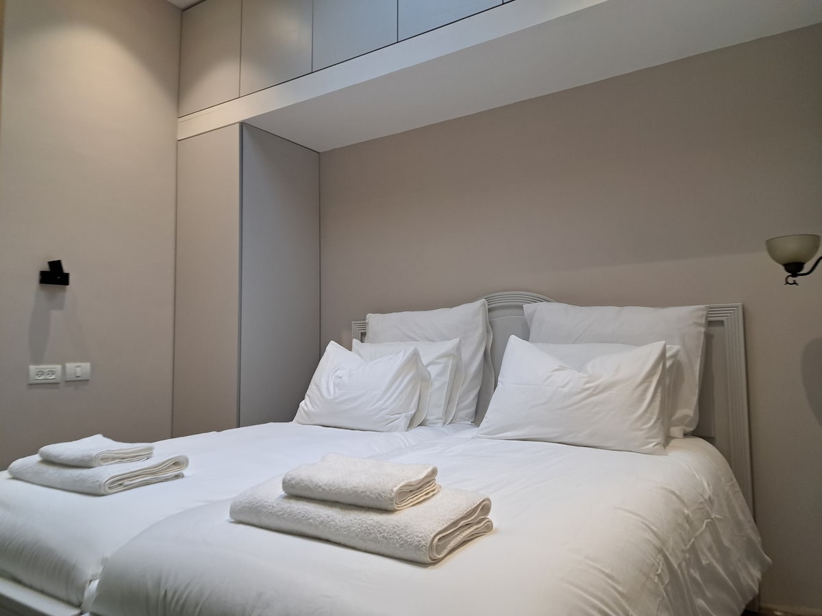 A comfortable sleeping area is presented with two spacious beds positioned side by side. Fresh white linens and neatly folded towels are arranged at the foot of each bed. Soft lighting enhances the neutral wall color, creating a calm and relaxing atmosphere.
