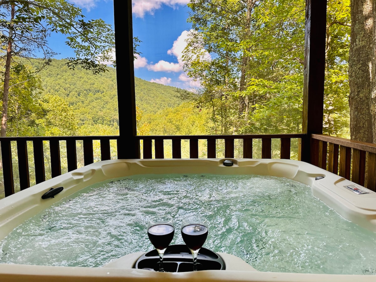 A serene hot tub sits on a screened deck, surrounded by lush greenery and framed by tall trees. Two glasses of red wine are placed on the edge of the tub, with panoramic mountain views visible through the mesh enclosure.