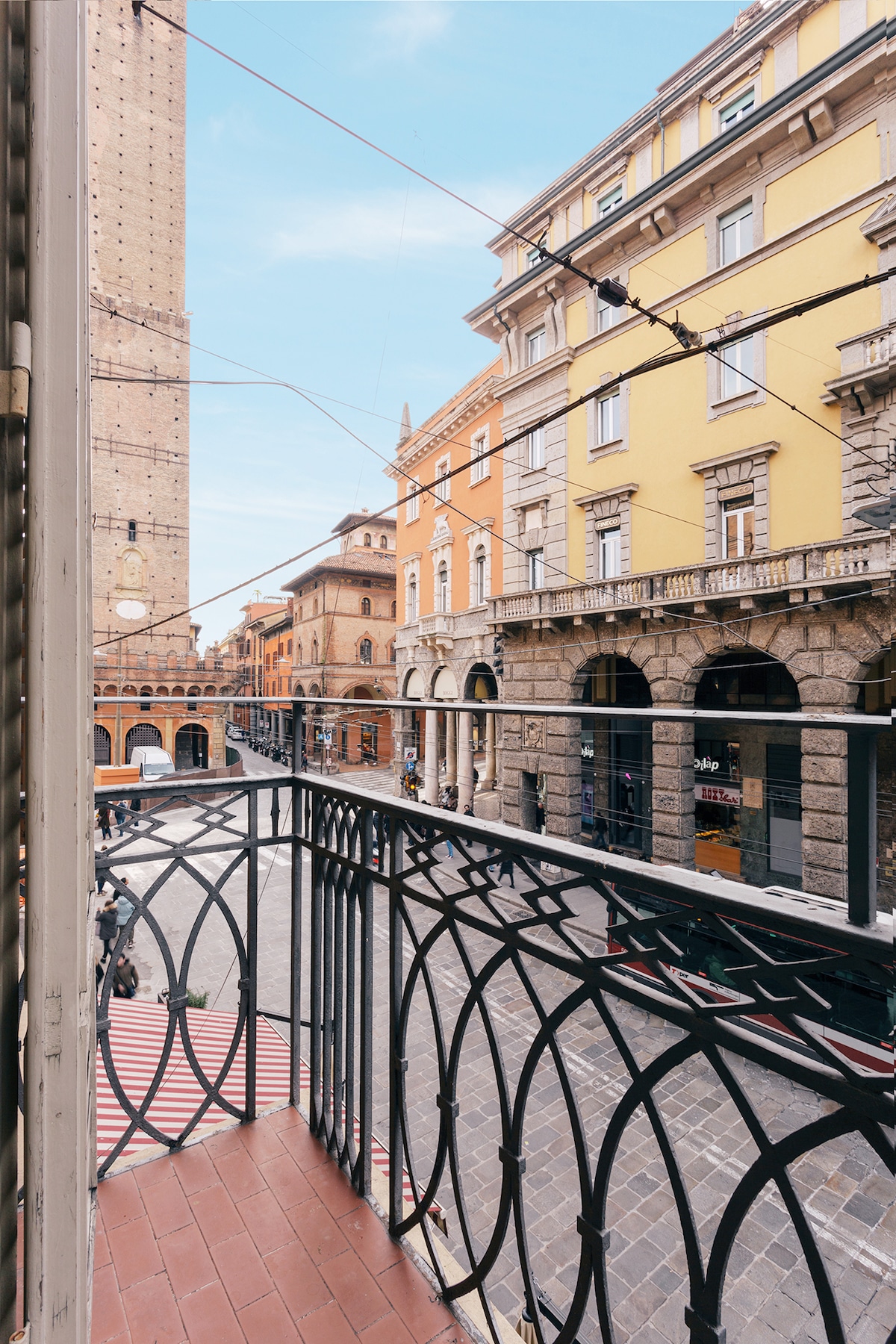 A decorative balcony with an intricate black railing is visible. Below, the vibrant street scene includes historic buildings in warm tones and the iconic towers of Bologna. A clear sky adds brightness to the view, enhancing the surrounding architecture.