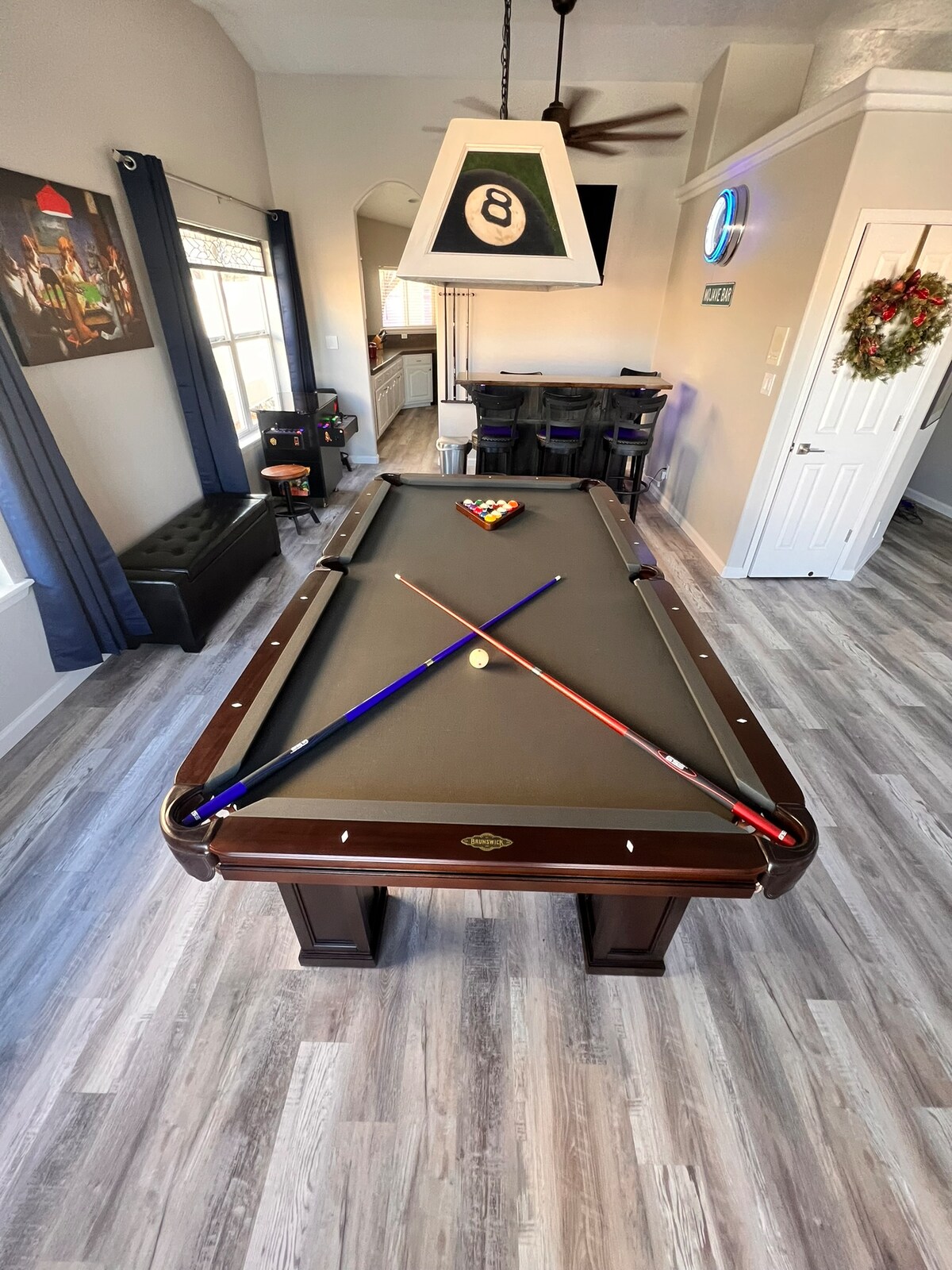 A Brunswick pool table is centered in the room, with colorful cues positioned diagonally across the surface. The space features modern furnishings, including a bar with tall chairs in the background and a pendant light overhead. Large windows allow natural light to illuminate the area.