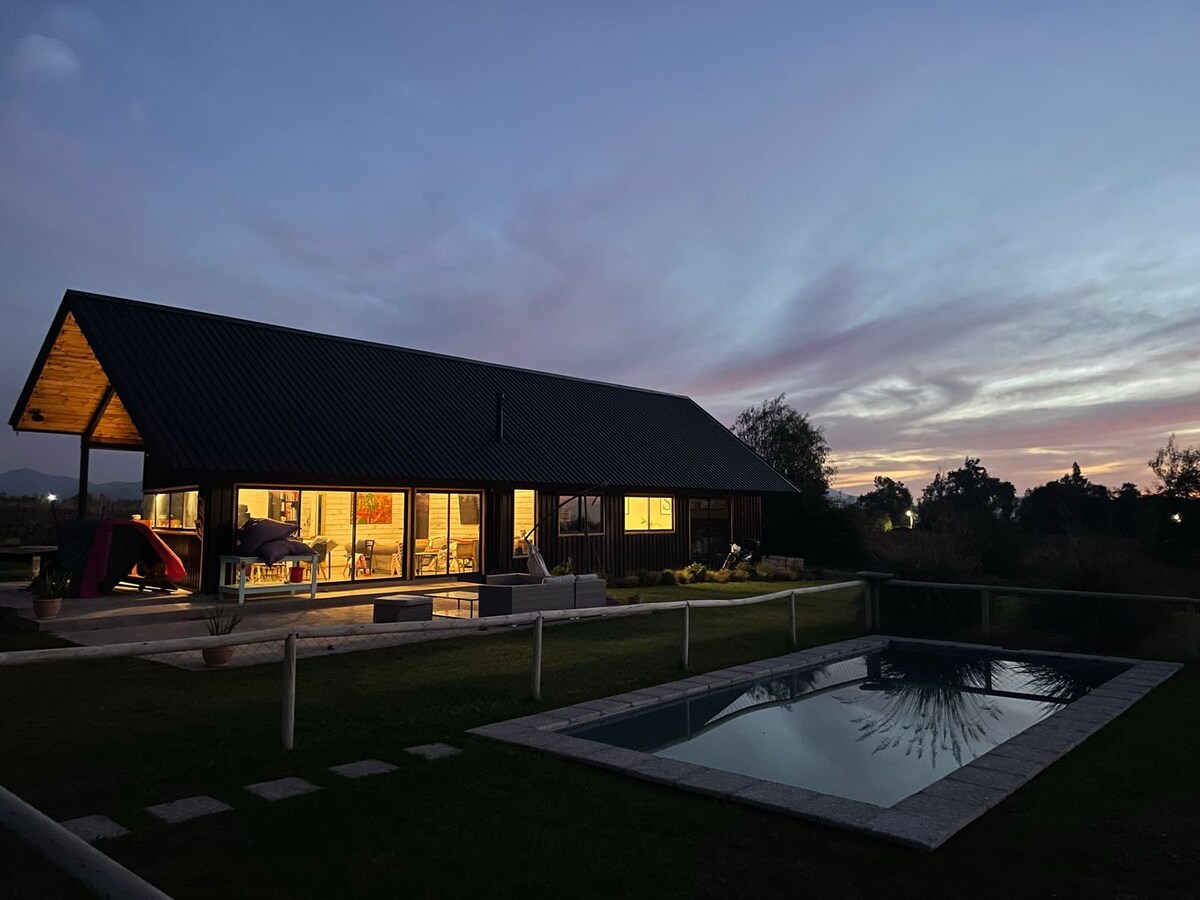 A spacious house is depicted against a sunset sky, showcasing large glass windows that reflect warm interior lighting. A pool is visible in the foreground, complemented by minimal landscaping and outdoor furniture, inviting relaxation in the serene environment.