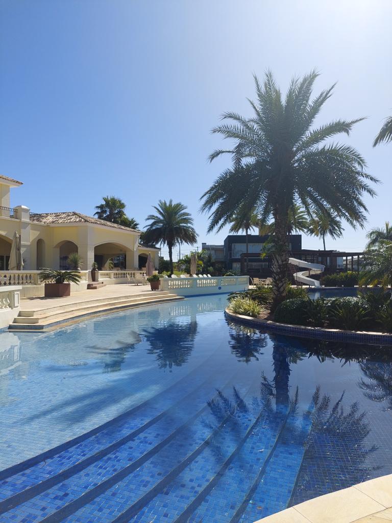 A large swimming pool with clear blue water reflects the surrounding palm trees and architecture. Steps lead to a spacious deck adorned with lush greenery. A water slide is visible in the background, offering an additional recreational feature.