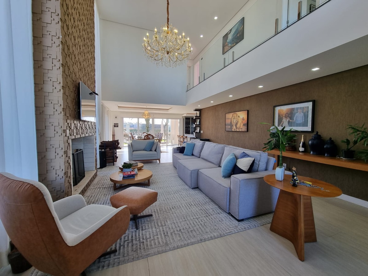 A spacious living area is featured, highlighted by a chandelier and large windows that allow natural light. A comfortable sectional sofa faces a TV mounted above the fireplace. A round coffee table and two accent chairs add to the inviting atmosphere.