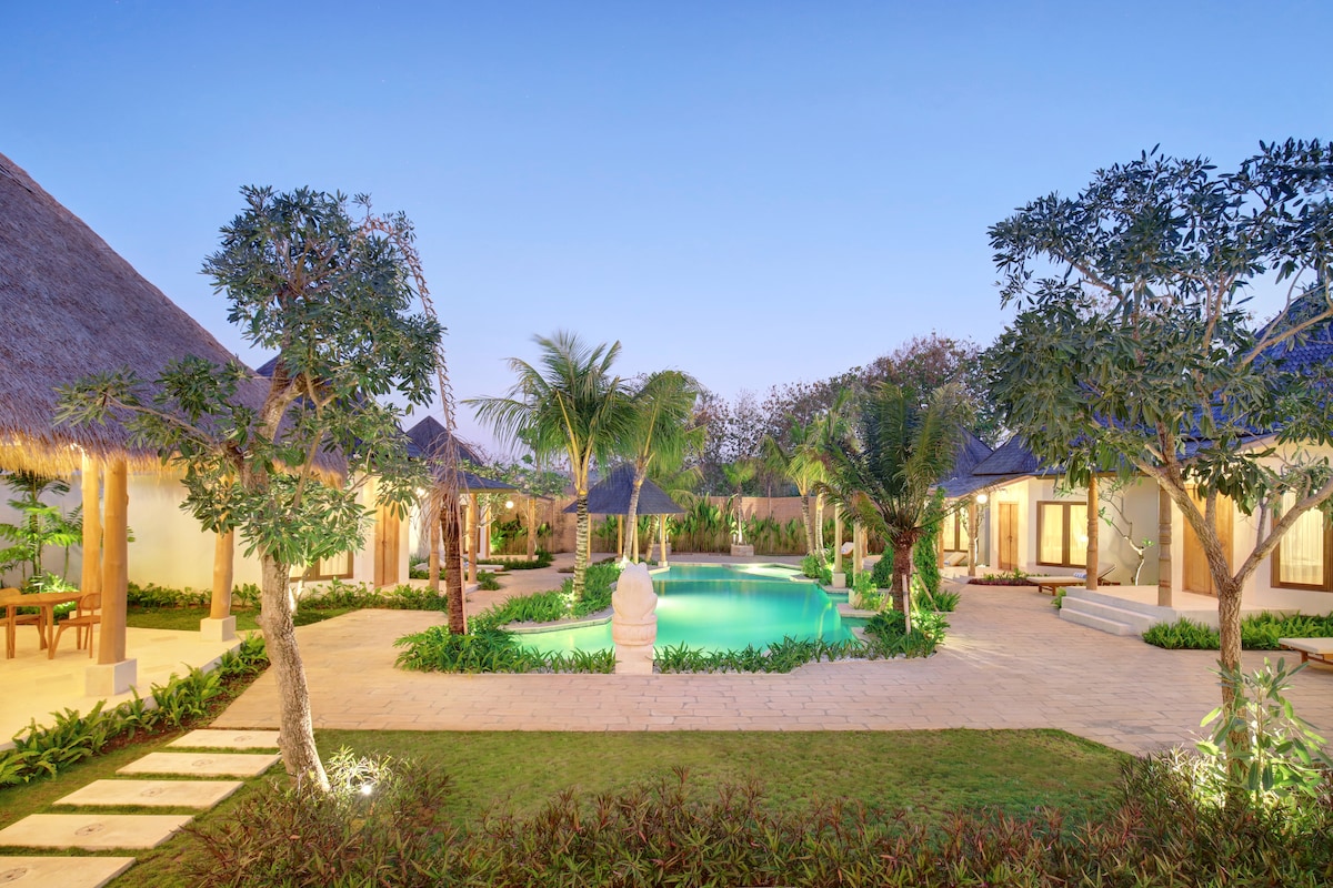 The image captures a serene outdoor area featuring a swimming pool surrounded by lush greenery and bungalow-style structures. Palm trees and garden plants add to the natural ambiance. Soft lighting enhances the pathways, creating a tranquil setting at dusk.