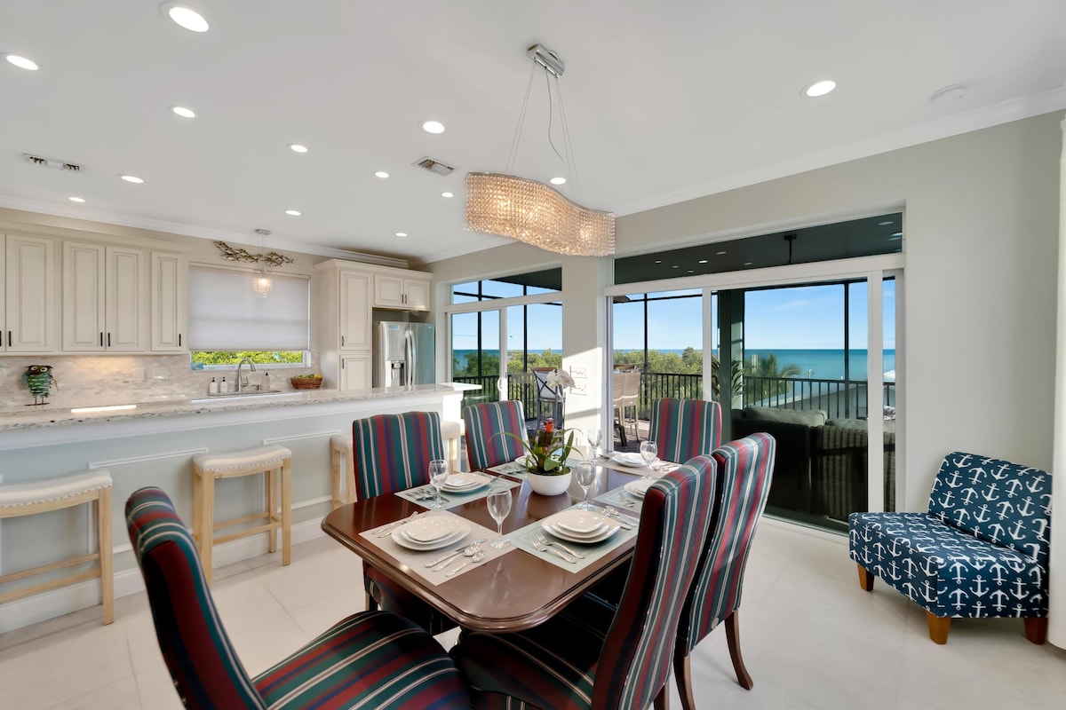 A dining area is highlighted by a sleek wooden table surrounded by striped chairs. Large windows offer views of the ocean, while a modern chandelier enhances the space. The kitchen area features light cabinetry and stainless steel appliances, creating a bright and open atmosphere.