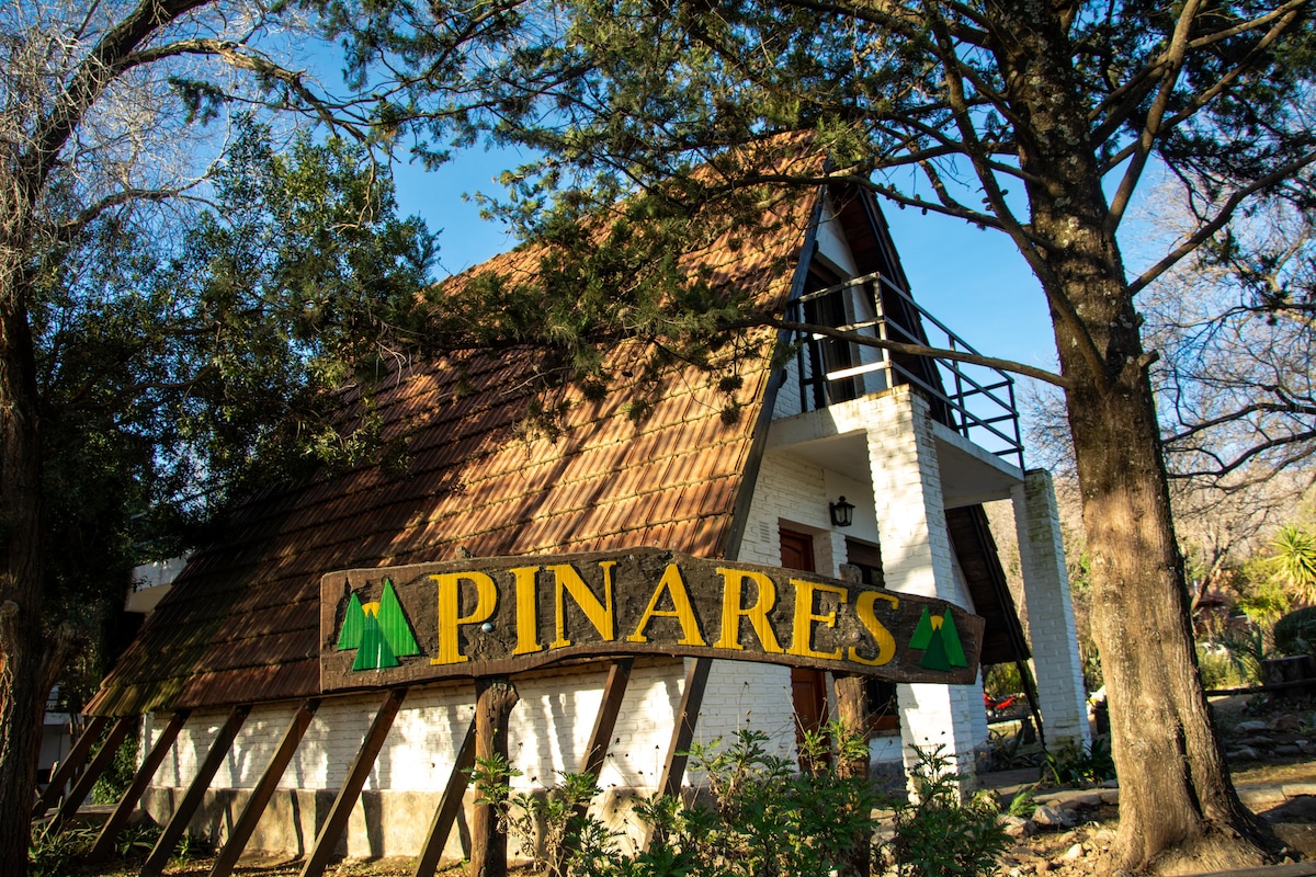 A charming triangular-roofed structure is framed by tall trees, with a wooden sign reading 'Pinares' prominently displayed in front. Natural light enhances the warm tones of the building's exterior, inviting guests to discover the peaceful surroundings.