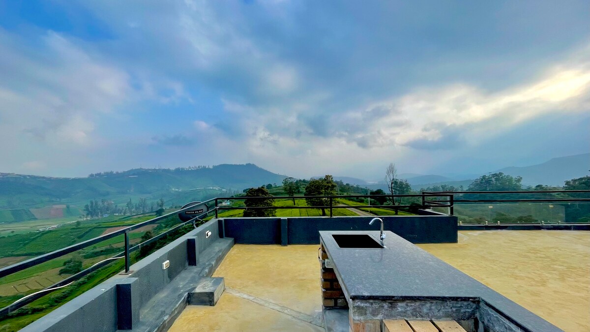 A rooftop bar area is showcased with a stone countertop and a sink. Surrounding views encompass rolling hills and lush greenery, set against a cloudy sky. A railing defines the space, enhancing the open-air atmosphere.