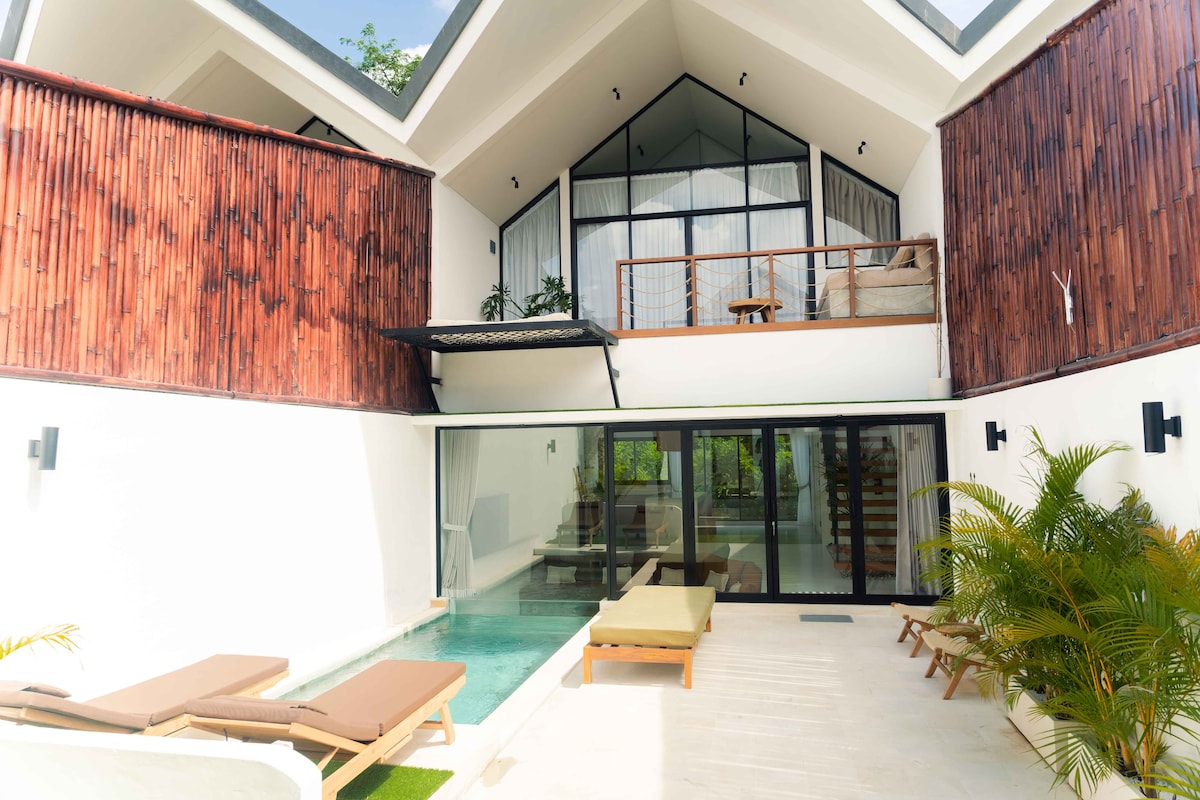 The villa features a modern architectural design with a spacious outdoor area, including a private pool. Two lounge chairs are positioned beside the pool, and an inviting seating area is visible on the upper balcony, surrounded by natural greenery.